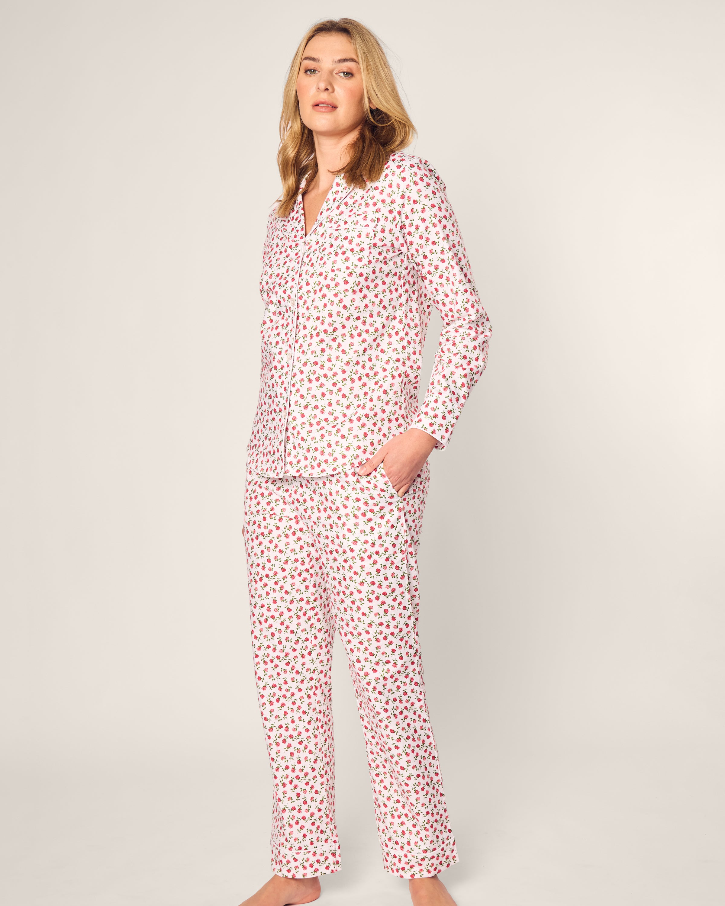 A person wears the Womens Twill Pajama Set in Petite Petals by Petite Plume, featuring pink and red florals on a light background. They pose with one hand in a pocket, showcasing luxurious sleepwear against a plain, light backdrop.
