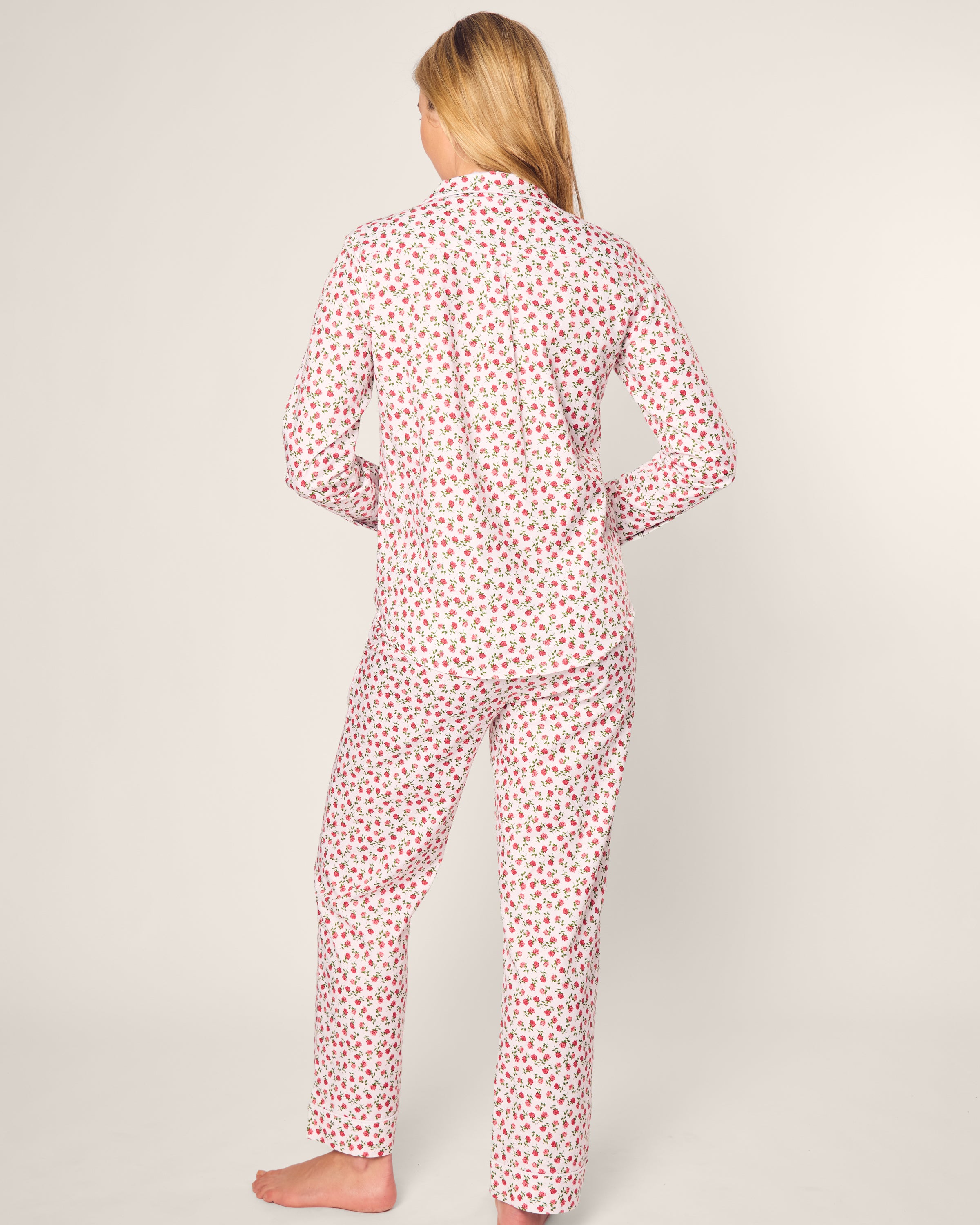 A person with long blonde hair stands barefoot, facing away, in Petite Plumes Womens Twill Pajama Set adorned with petite red heart petals on a white base. The luxurious sleepwear features a matching long-sleeve top and pants. The background is plain beige.