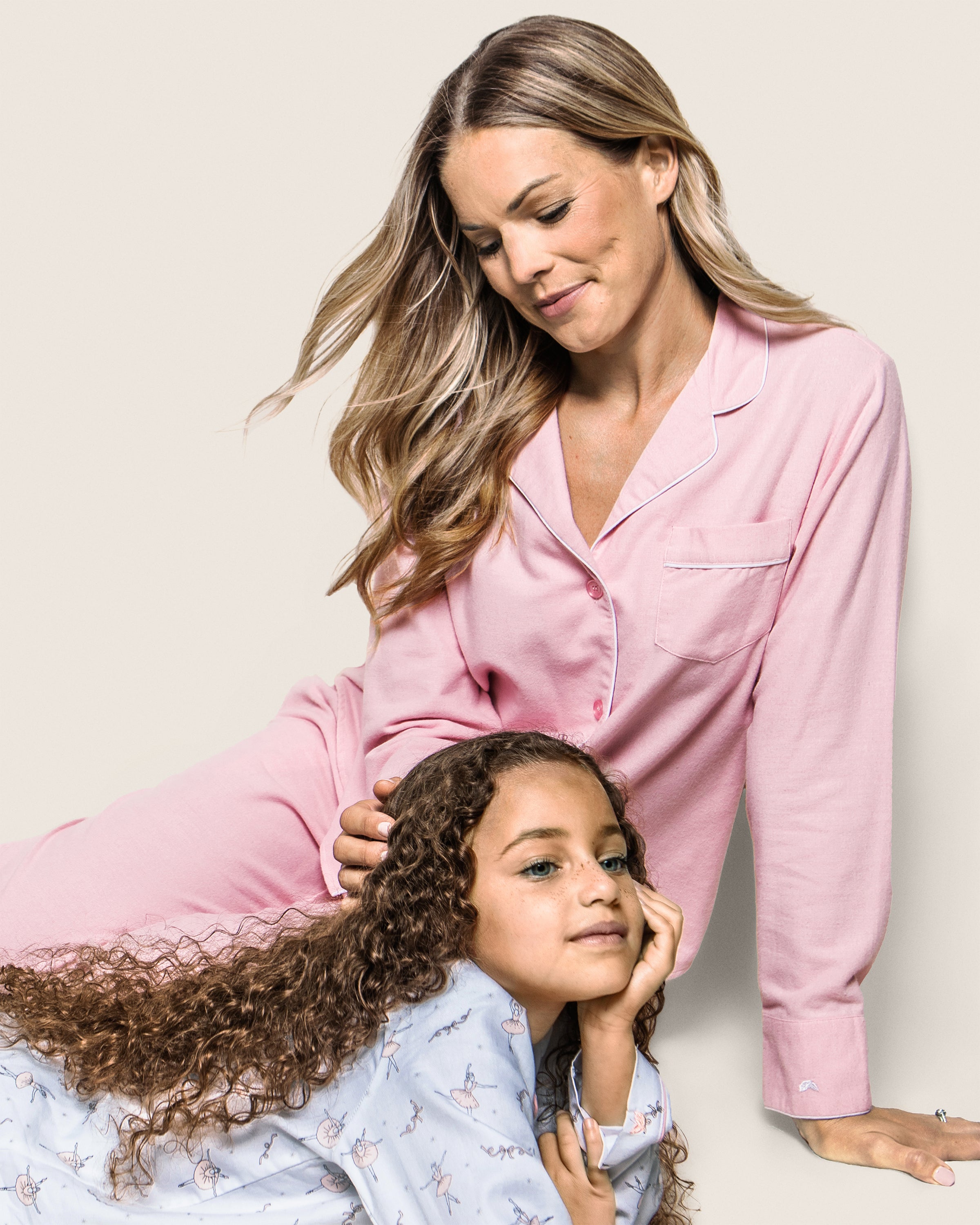 A woman in a Petite Plume Womens Flannel Pajama Set in Pink gently holds a childs head, who is wearing blue dinosaur pajamas. Both have relaxed expressions against a soft, neutral background.