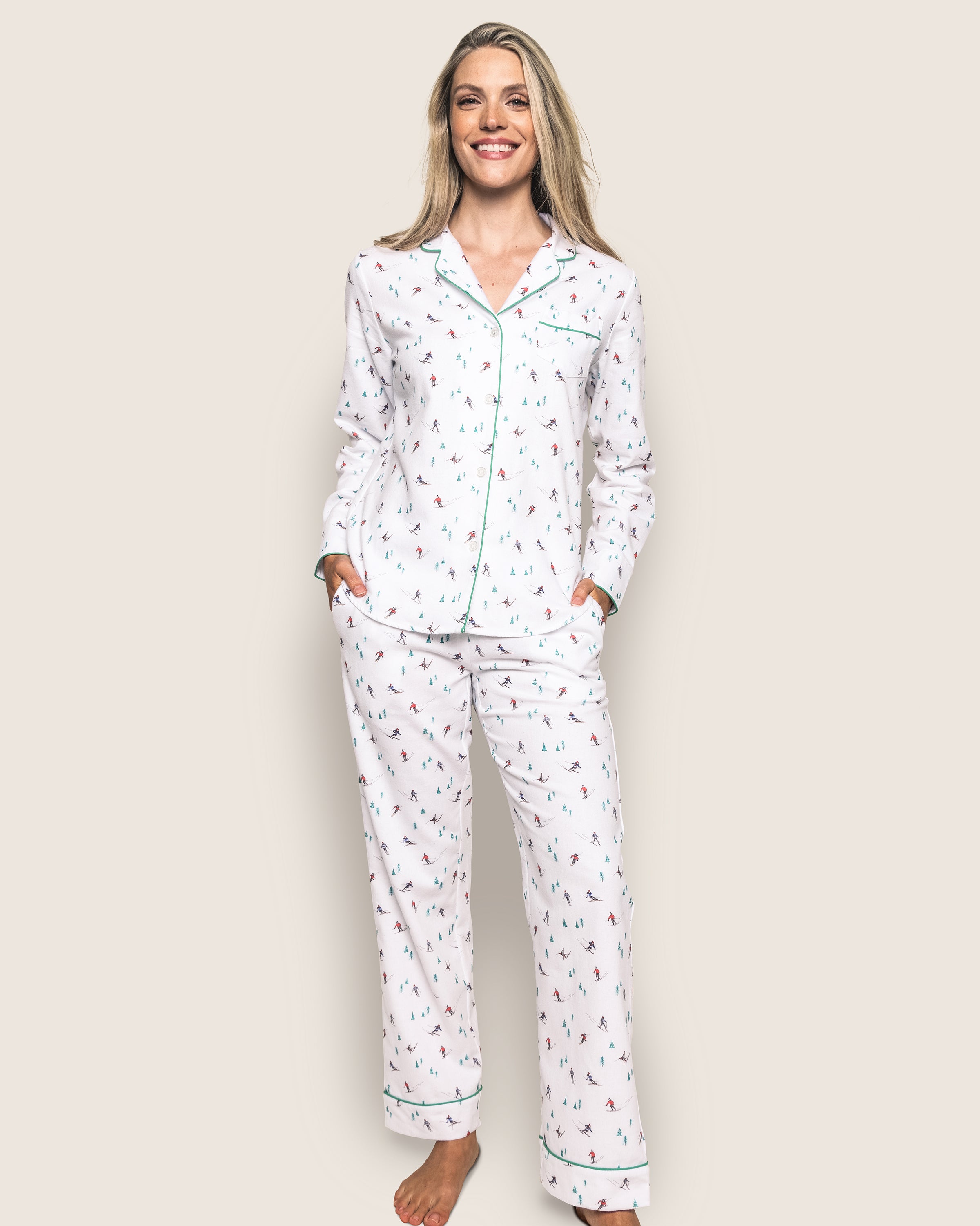 A person stands smiling, wearing the luxurious Womens Flannel Pajama Set in Après Ski by Petite Plume. The top features green piping and a collar, while hands are in the pockets of loose-fitting, soft brushed cotton flannel pants. The background is a plain light color.
