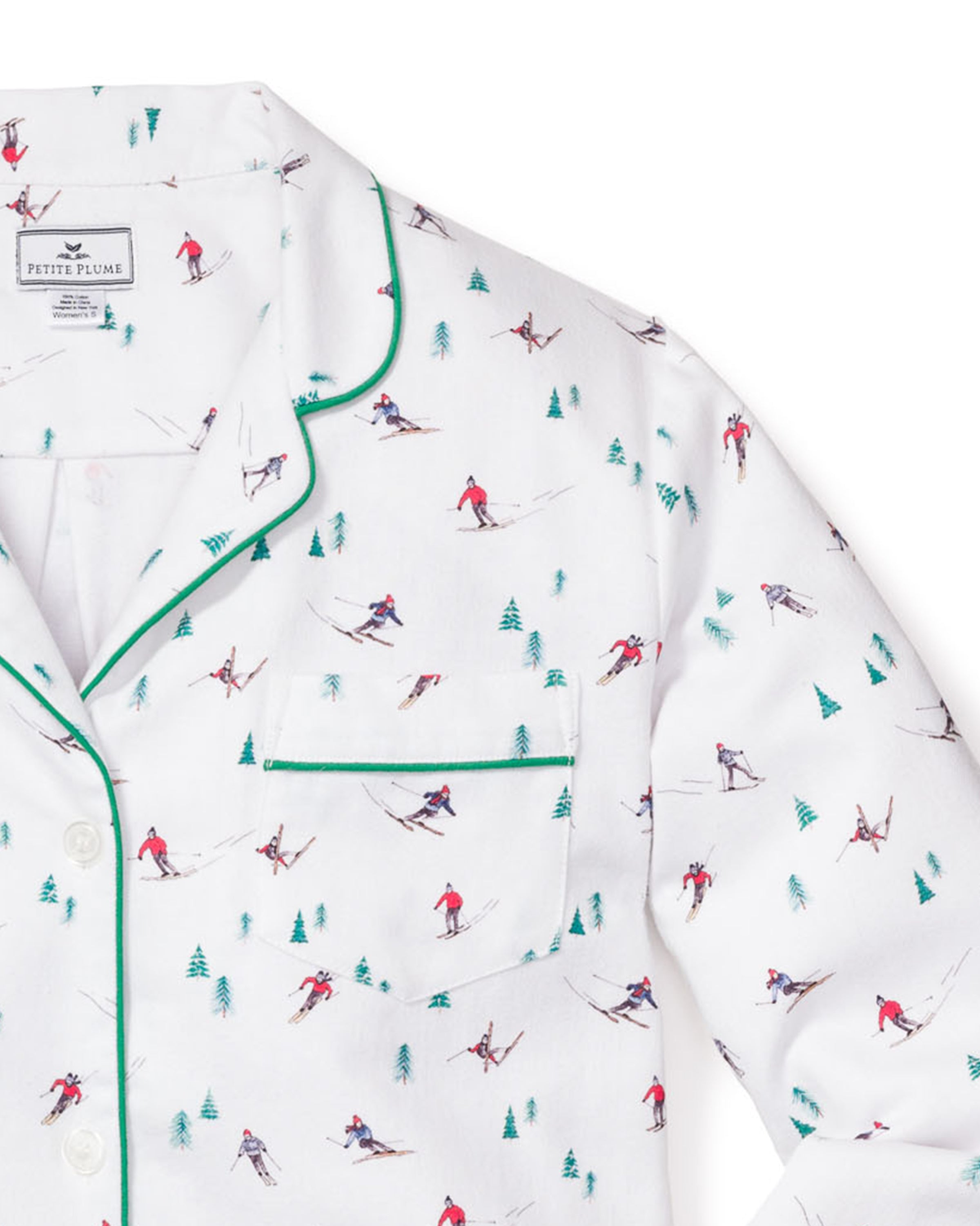The Womens Flannel Pajama Set in Après Ski by Petite Plume is a white brushed cotton flannel top featuring a vibrant skier and pine tree pattern, accented with green piping on the collar and pocket for stylish comfort.