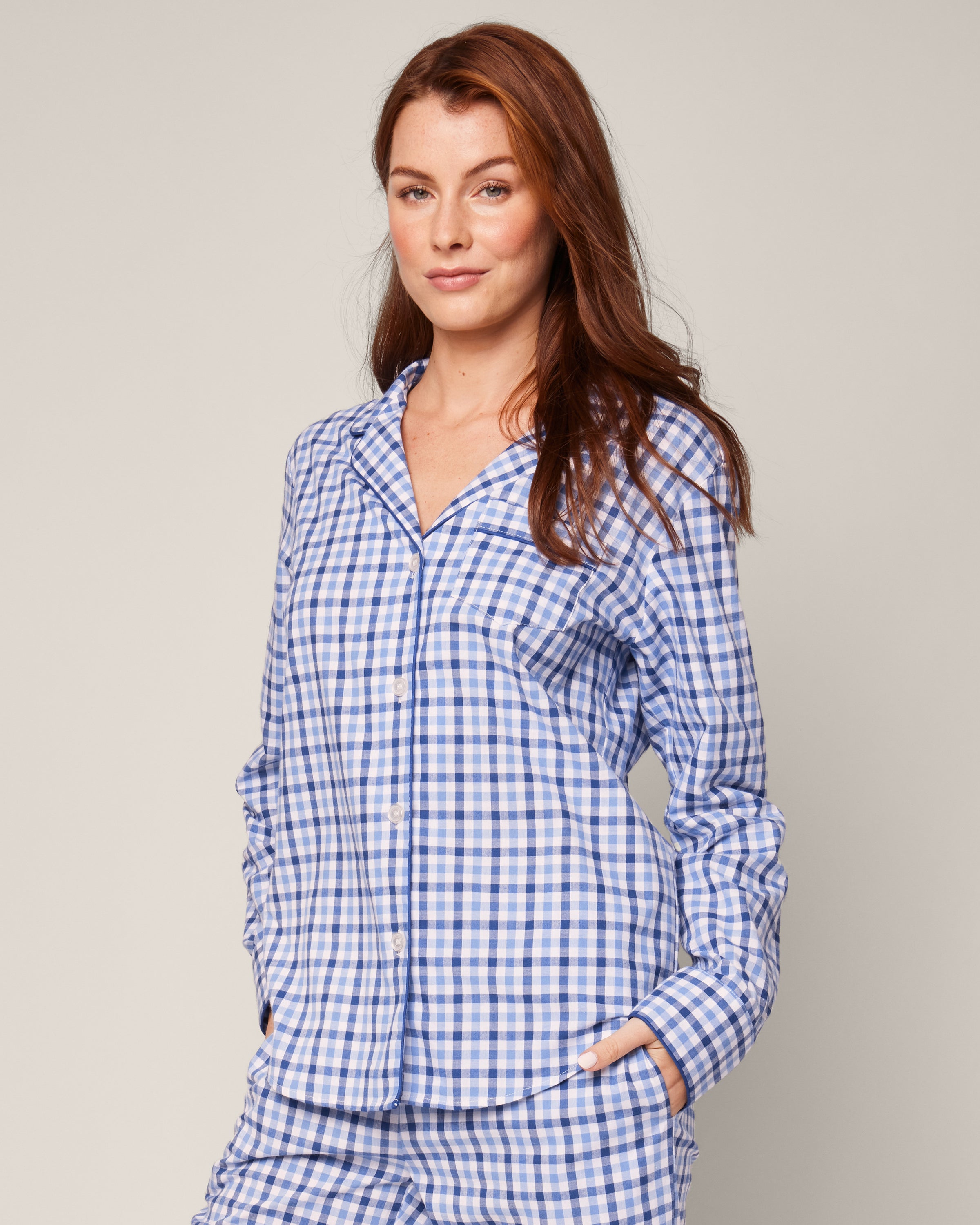 A person with long auburn hair is wearing the Womens Twill Pajama Set in Royal Blue Gingham by Petite Plume, featuring a collared shirt with button closure, standing with one hand in their pocket against a plain light gray background.