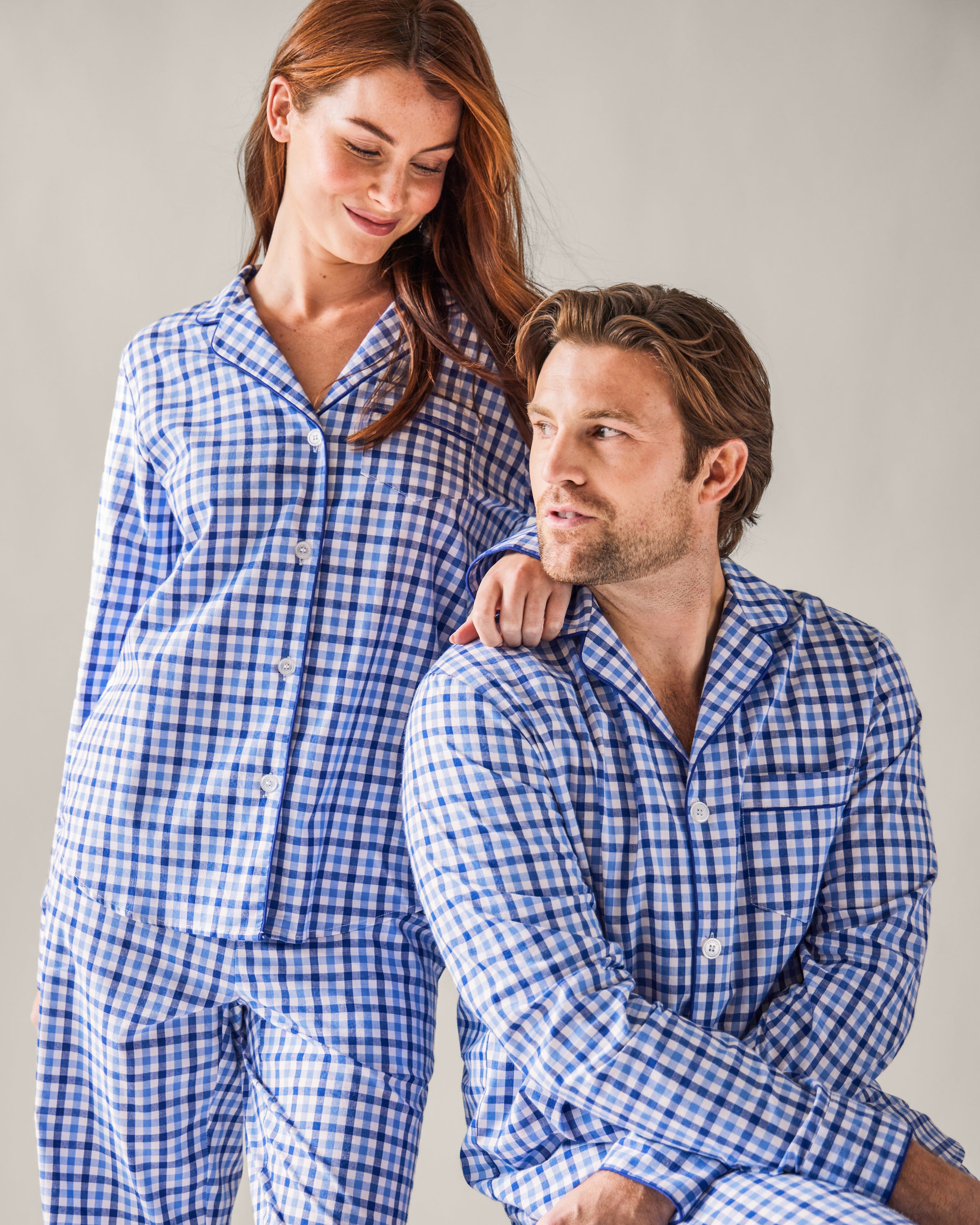 A woman and man in matching blue and white gingham pajamas embody the luxury of Petite Plume. She wears the Womens Twill Pajama Set in Royal Blue Gingham and smiles, placing her hand on his shoulder while he gazes up at her against a neutral background.