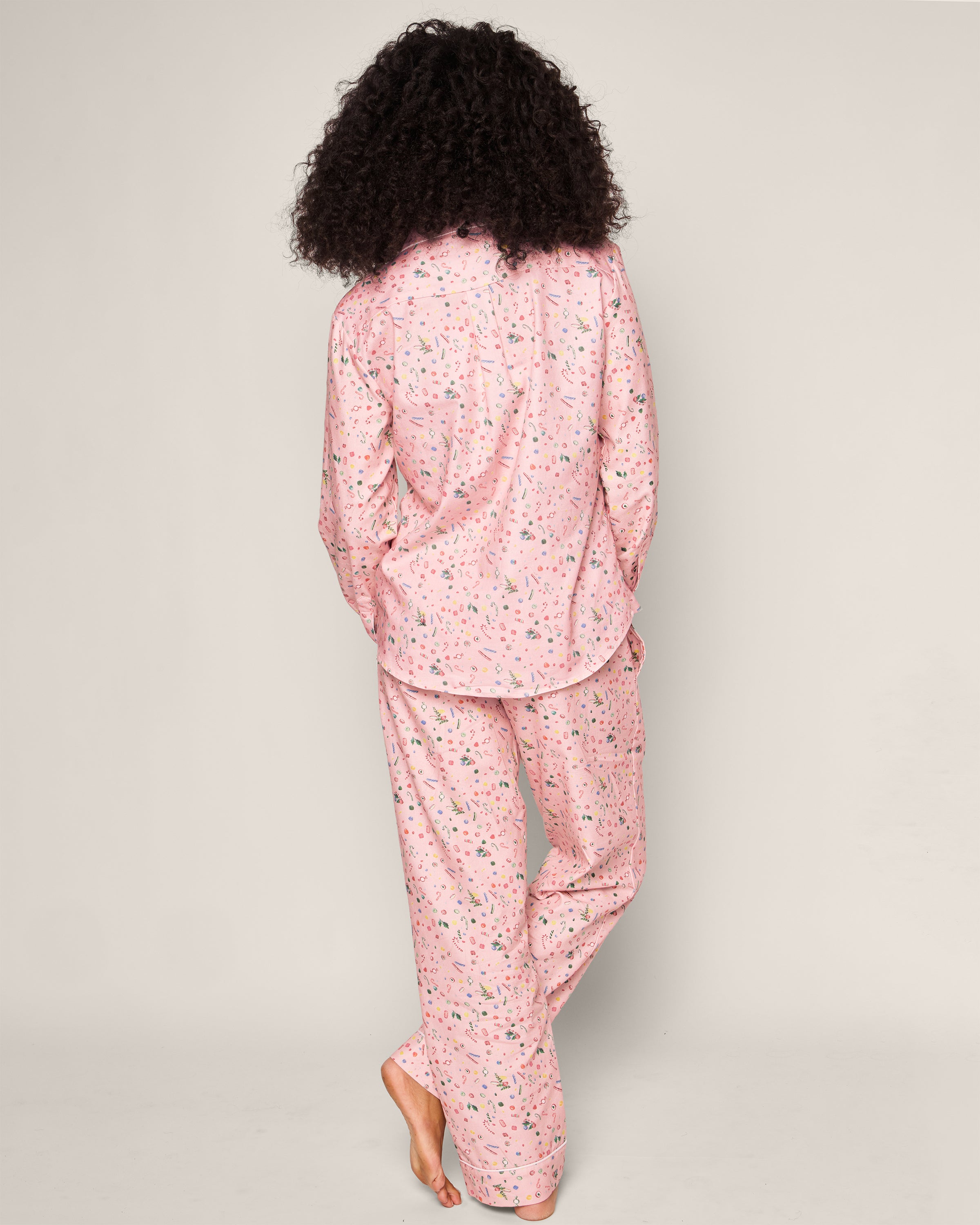 A barefoot person with curly hair poses against a plain backdrop, wearing Petite Plumes Womens Twill Pajama Set in Vintage Sweets, featuring nostalgic floral pink pajamas.