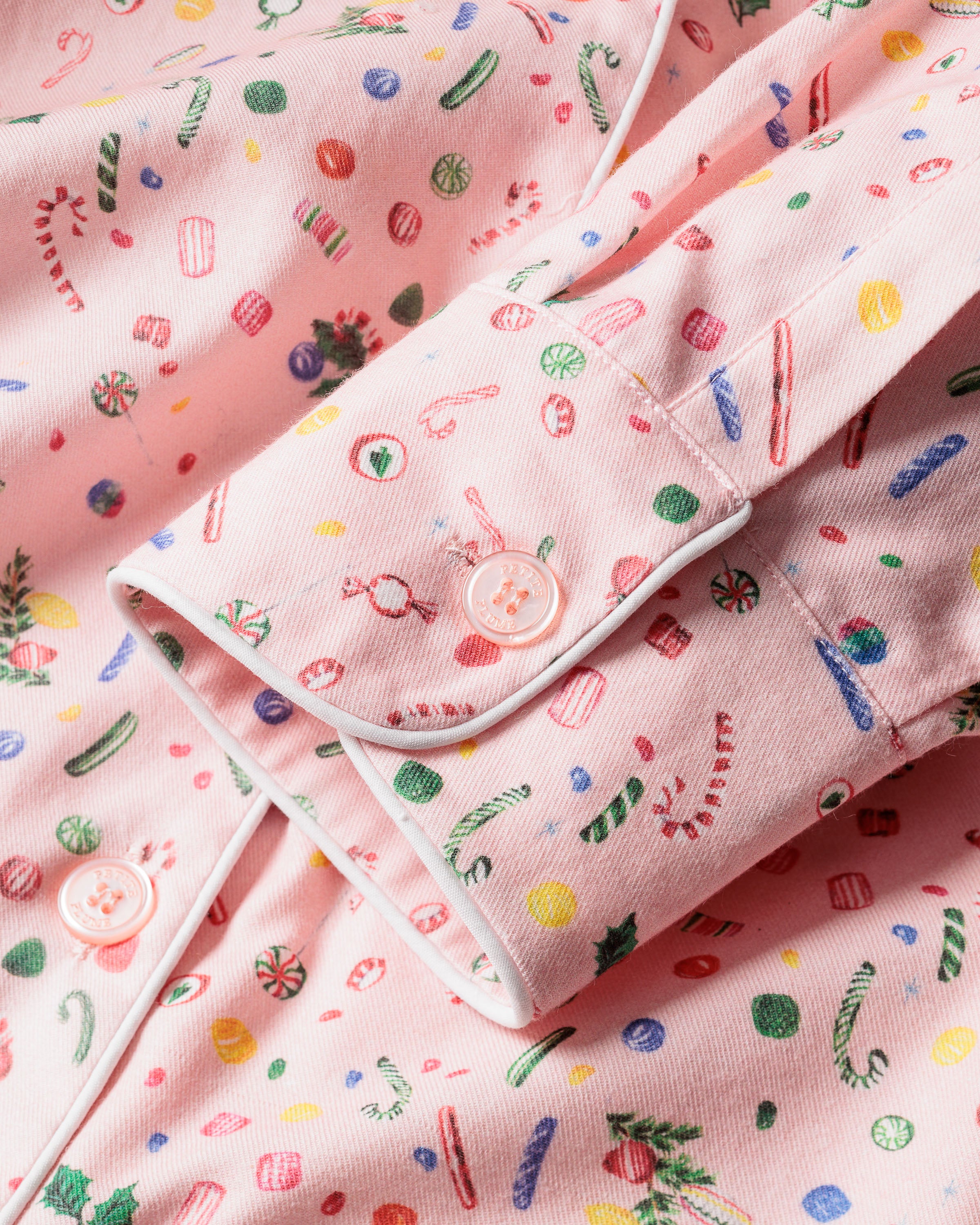 Close-up of Petite Plumes Womens Twill Pajama Set in Vintage Sweets, showcasing a pink fabric with candy canes, lollipops, and candies. The shirt sleeve has white piping and pink buttons, echoing holiday sweetness.
