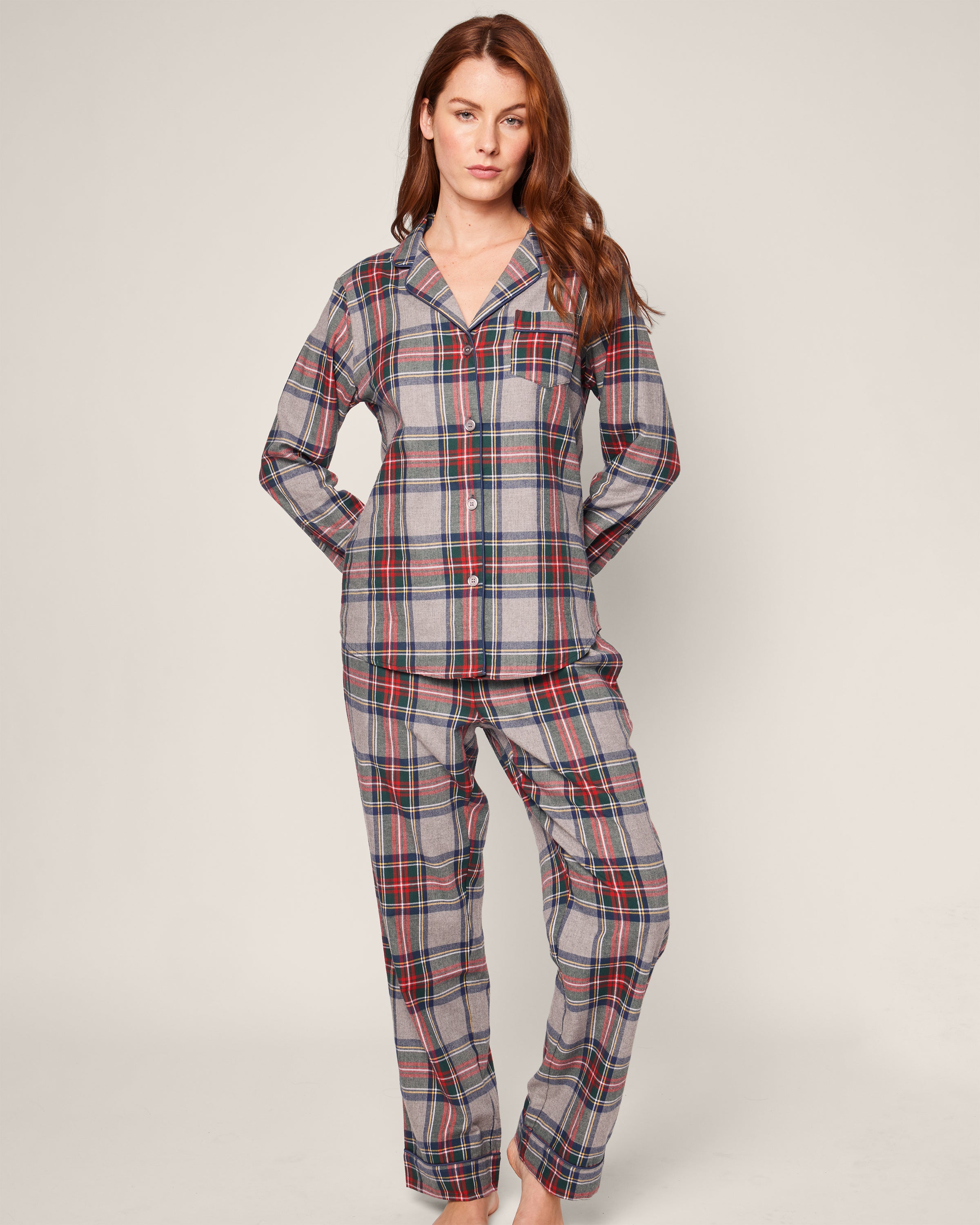 A person is wearing the Womens Brushed Cotton Pajama Set in Westminster Tartan by Petite Plume, featuring red, blue, and gray plaid patterns. This luxurious set includes a long-sleeved button-up shirt with a chest pocket and long pants against an off-white background.