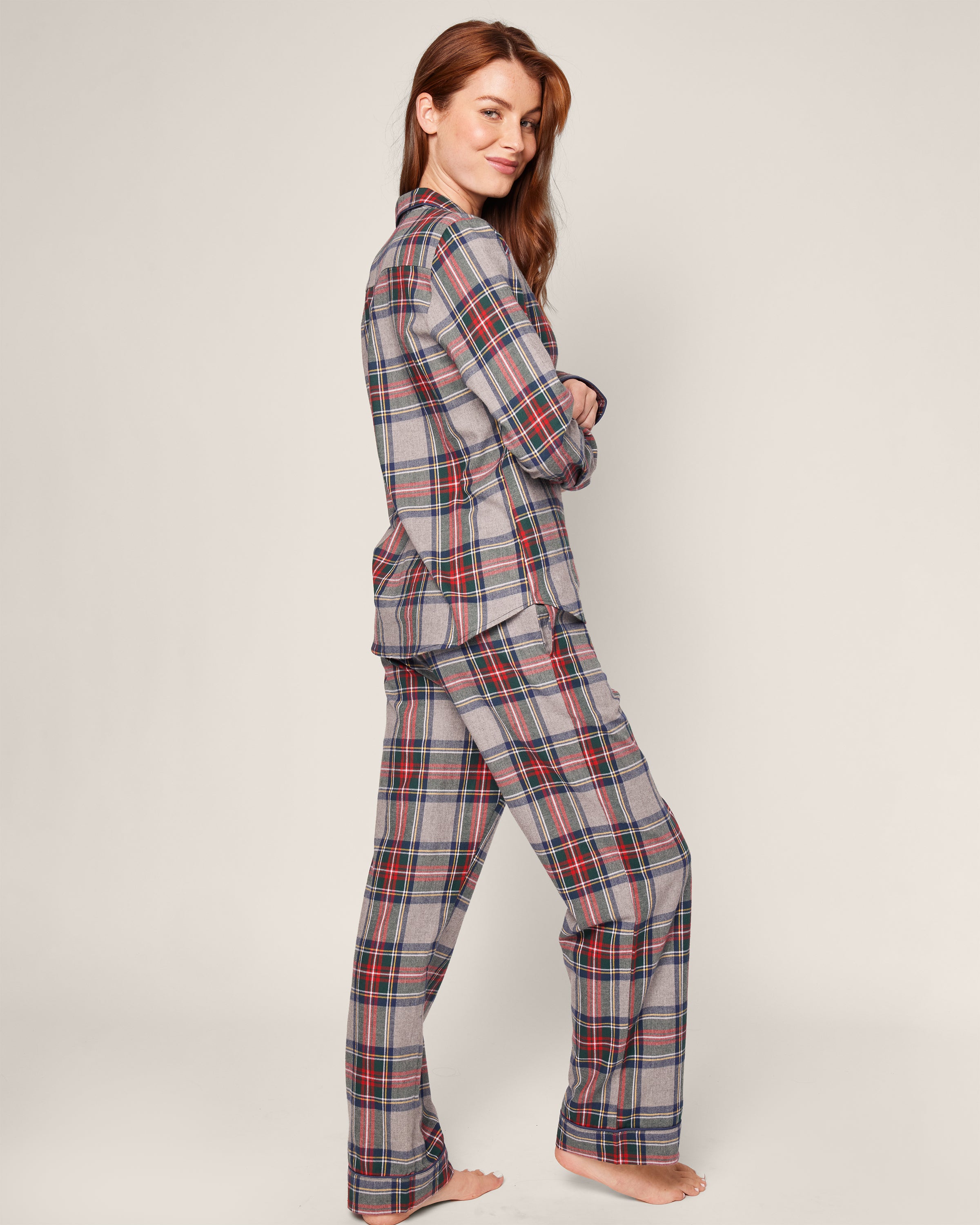 A smiling person stands barefoot, arms crossed, in Petite Plumes Womens Brushed Cotton Pajama Set with red, blue, and white Westminster Tartan patterns. The plain background enhances the cozy nod to Scottish tradition.