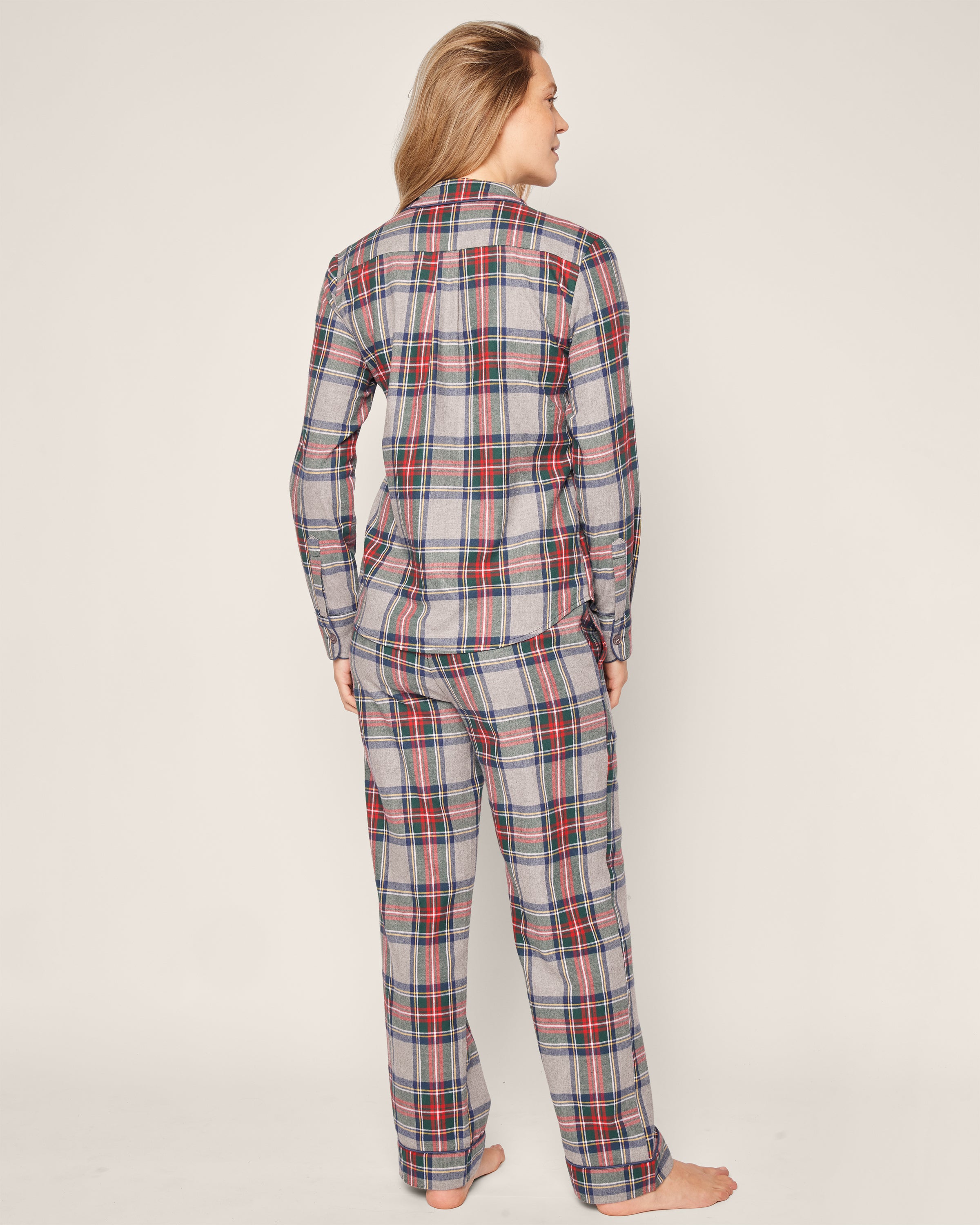 A person in a Petite Plume Womens Brushed Cotton Pajama Set, inspired by Westminster Tartan with a red, blue, and gray plaid pattern, stands barefoot against a plain background. They face slightly to the side with long hair cascading down their back.
