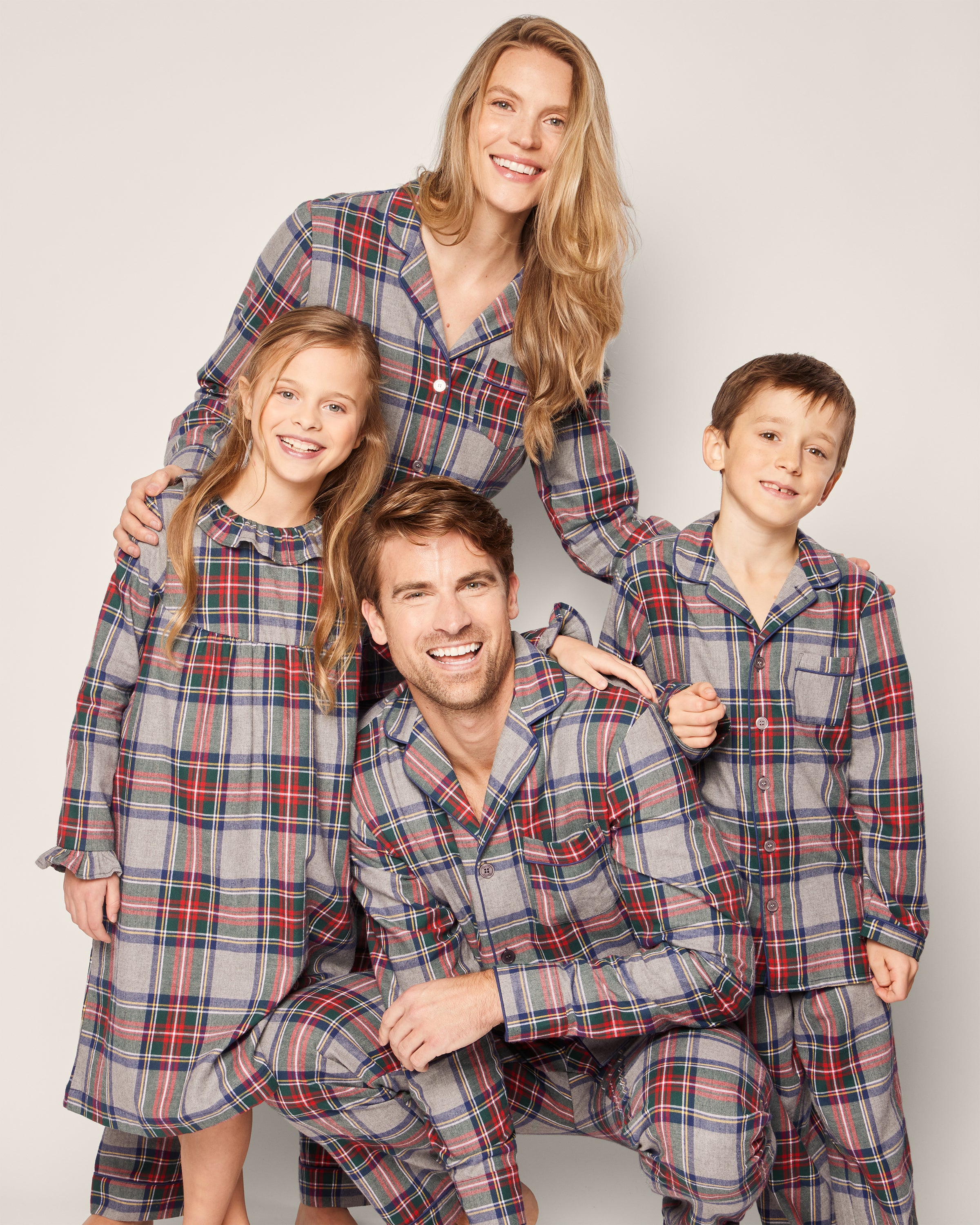A family of four, clad in Petite Plumes Womens Brushed Cotton Pajama Set in Westminster Tartan, shares a joyful moment. The parents stand while the children are beside them, all smiling against a plain backdrop that highlights the luxurious Scottish tradition of their sleepwear.