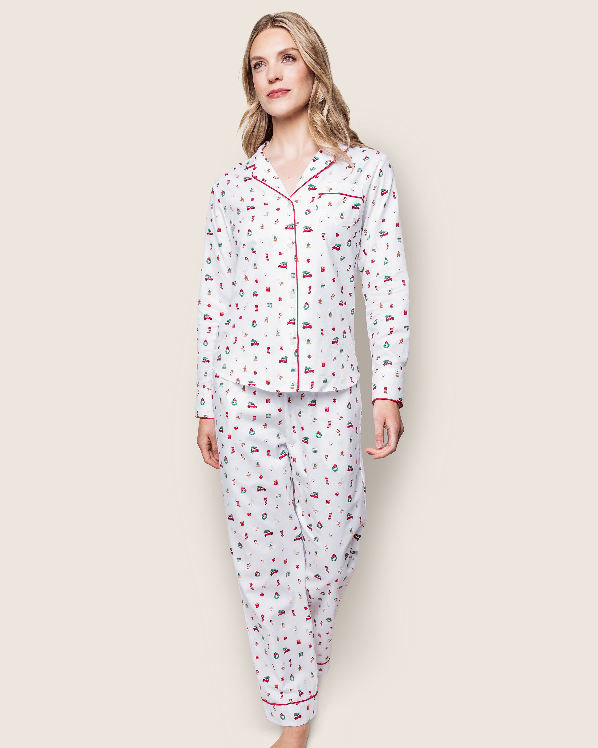 A person confidently walks wearing the Petite Plume Womens Twill Pajama Set in Winter Nostalgia. The white set features colorful illustrations, red piping on the cuffs and collar, a button-up top, and matching pants—perfect for the holiday season against a plain light background.