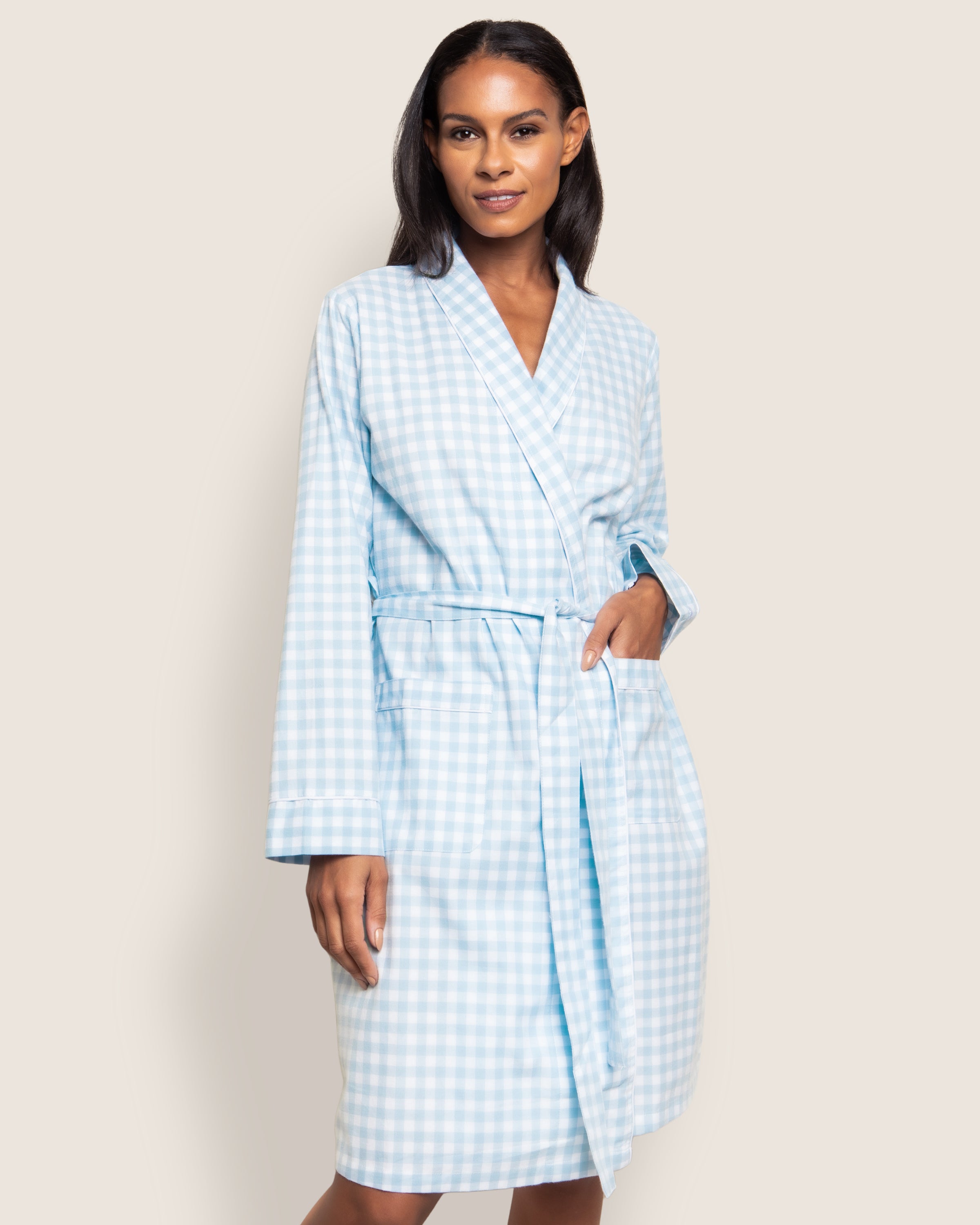 A person wears Petite Plumes Womens Twill Robe in Light Blue Gingham, crafted from high-quality cotton. The knee-length luxurious robe features a belt and long sleeves. They stand against a plain light backdrop, looking at the camera with a neutral expression.