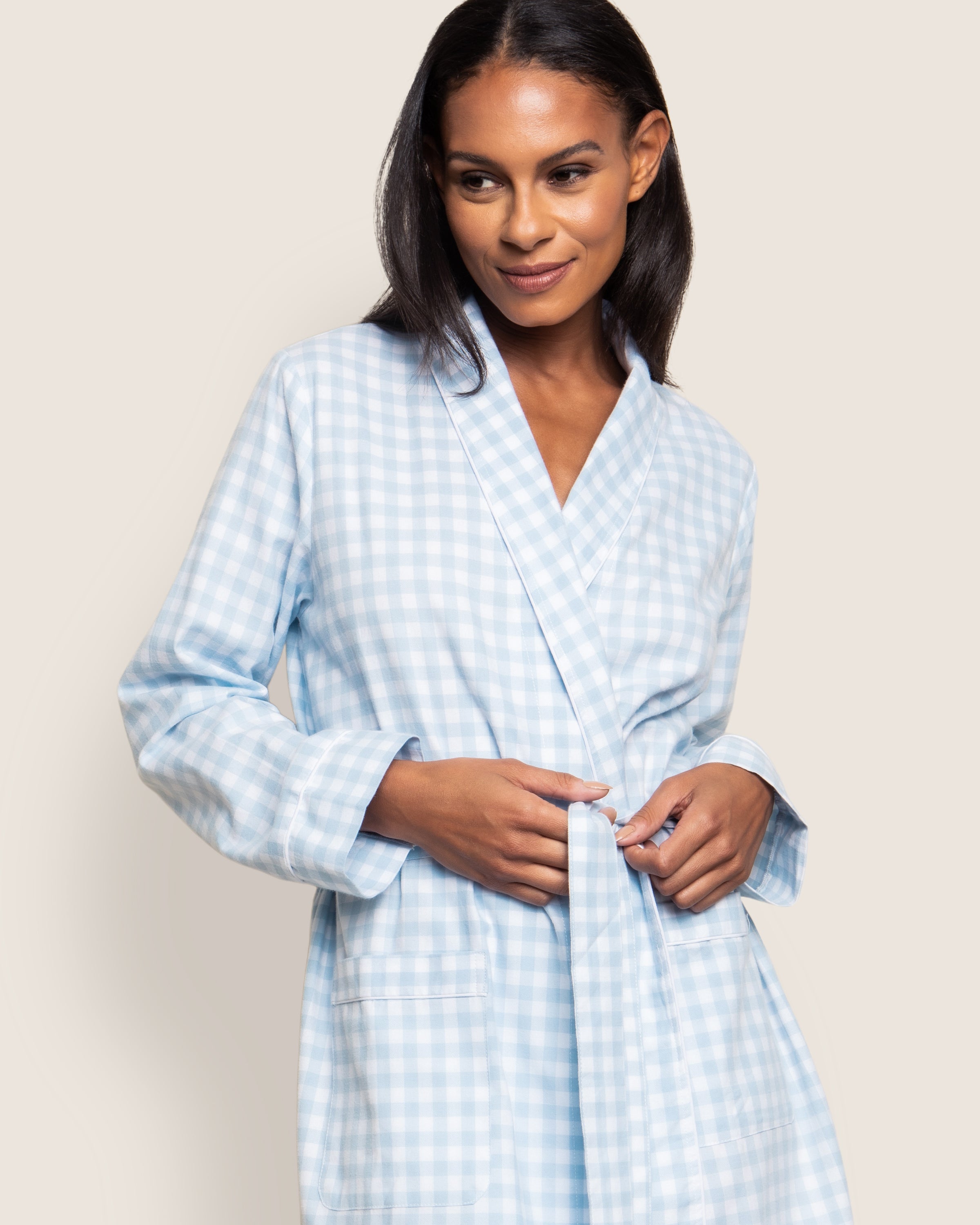 A person smiles while tying the belt of Petite Plumes Womens Twill Robe in Light Blue Gingham, made from premium cotton. The robe features long sleeves and two front pockets, showcasing luxurious sleepwear against a neutral backdrop.