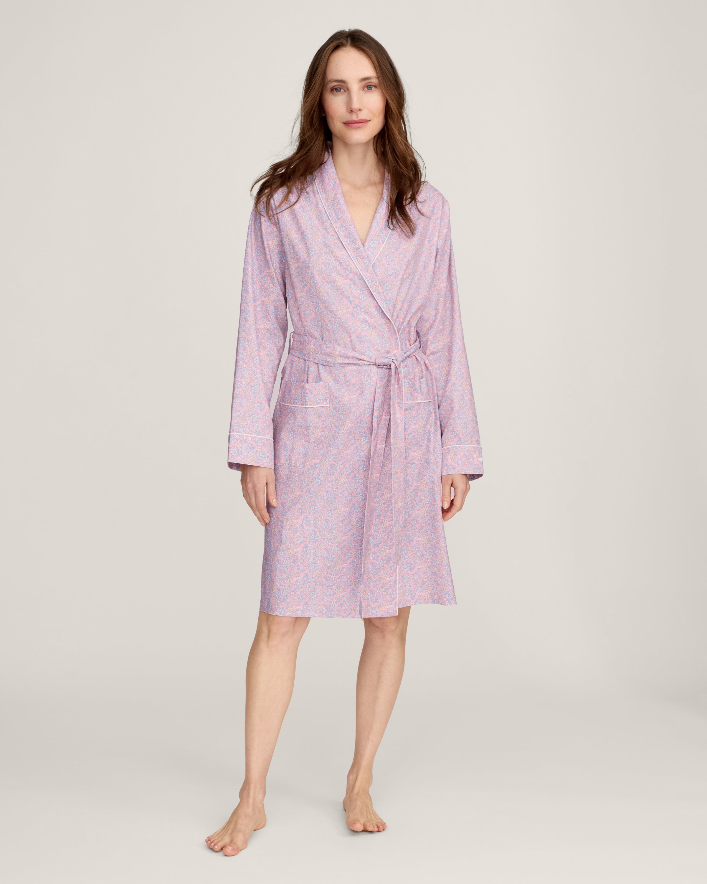 A woman stands barefoot against a plain light background, wearing the Women's Twill Robe in Fleur Blush—a knee-length, long-sleeved, light pink robe with a subtle floral pattern and waist tie.