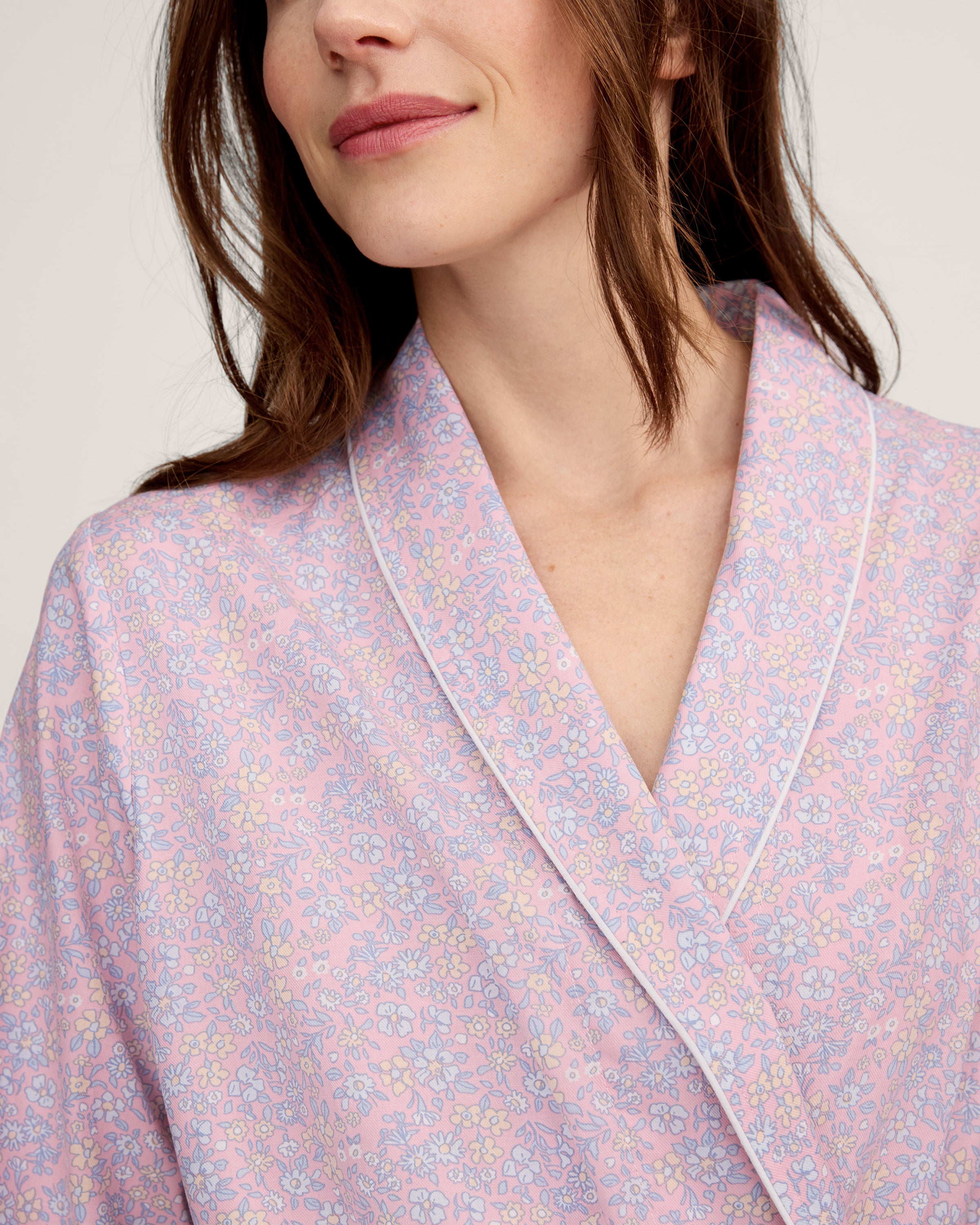 A woman stands barefoot against a plain light background, wearing the Women's Twill Robe in Fleur Blush—a knee-length, long-sleeved, light pink robe with a subtle floral pattern and waist tie.