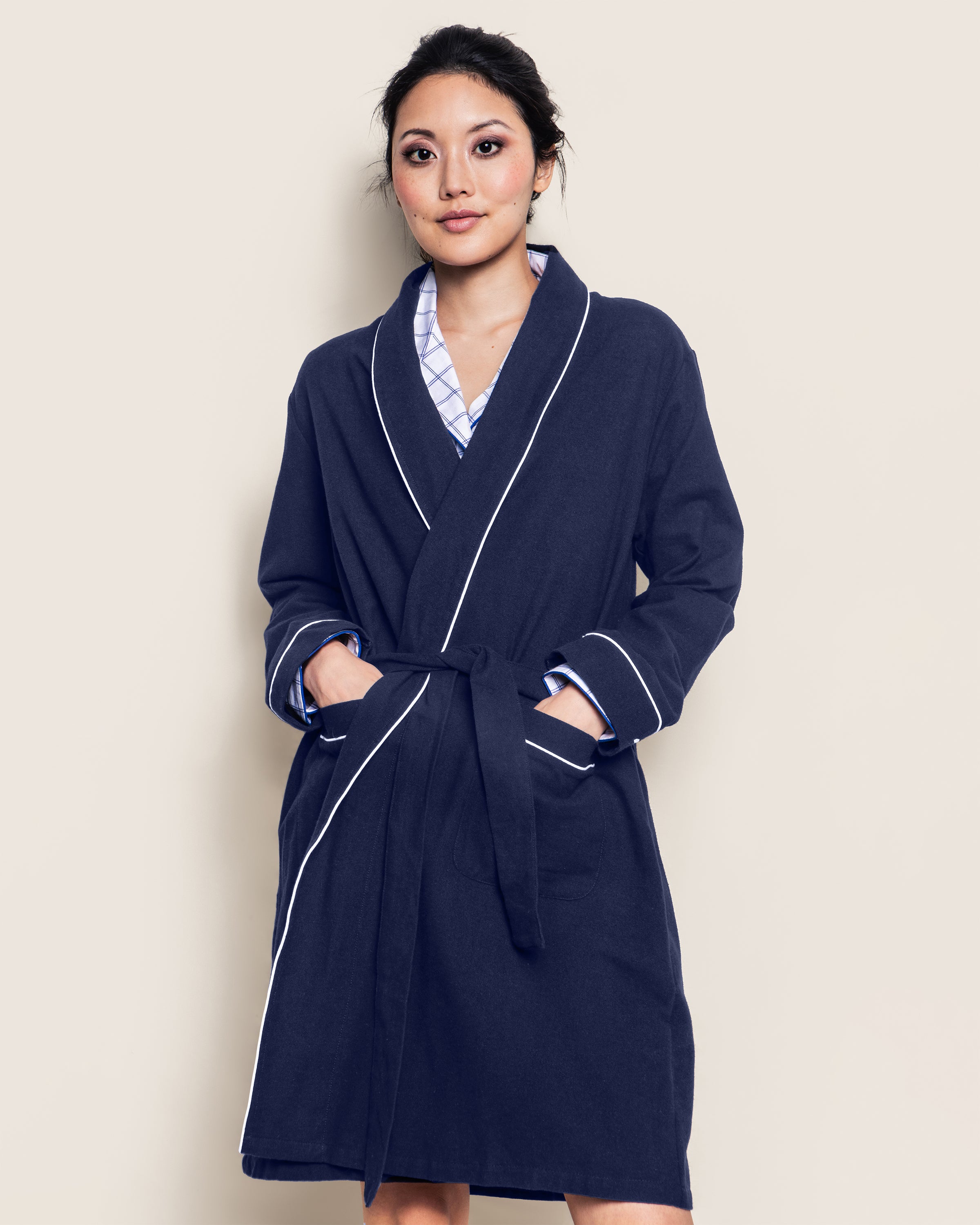 A person stands against a beige background, wearing the Womens Flannel Robe in Navy by Petite Plume. Made from 100% cotton twill, it features white trim and a waist belt. The person has their hands in the pockets, looking directly at the camera.