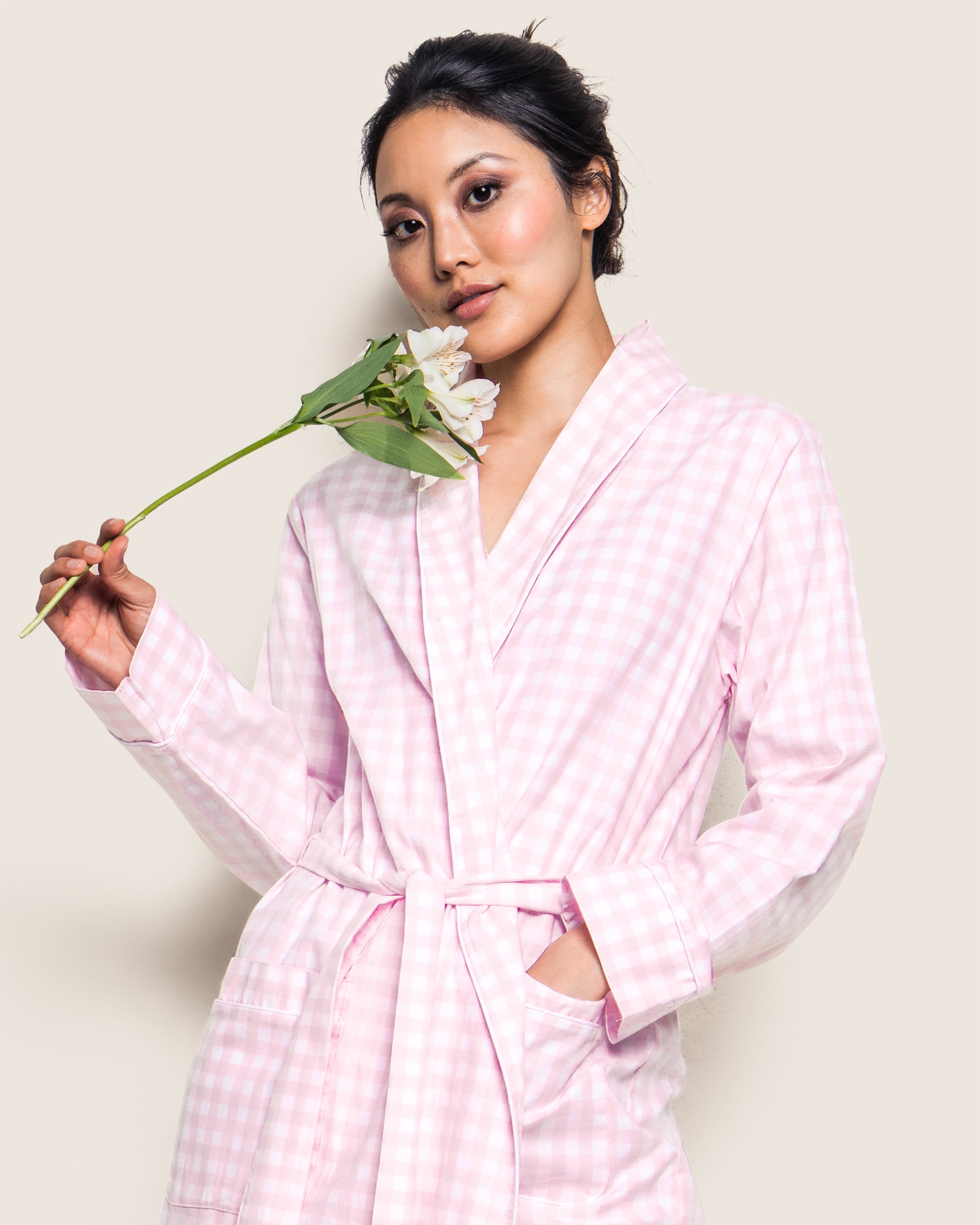A person in a Womens Twill Robe in Pink Gingham by Petite Plume holds a white flower near their face. With a relaxed expression and one hand in their pocket, they stand against a neutral beige background.