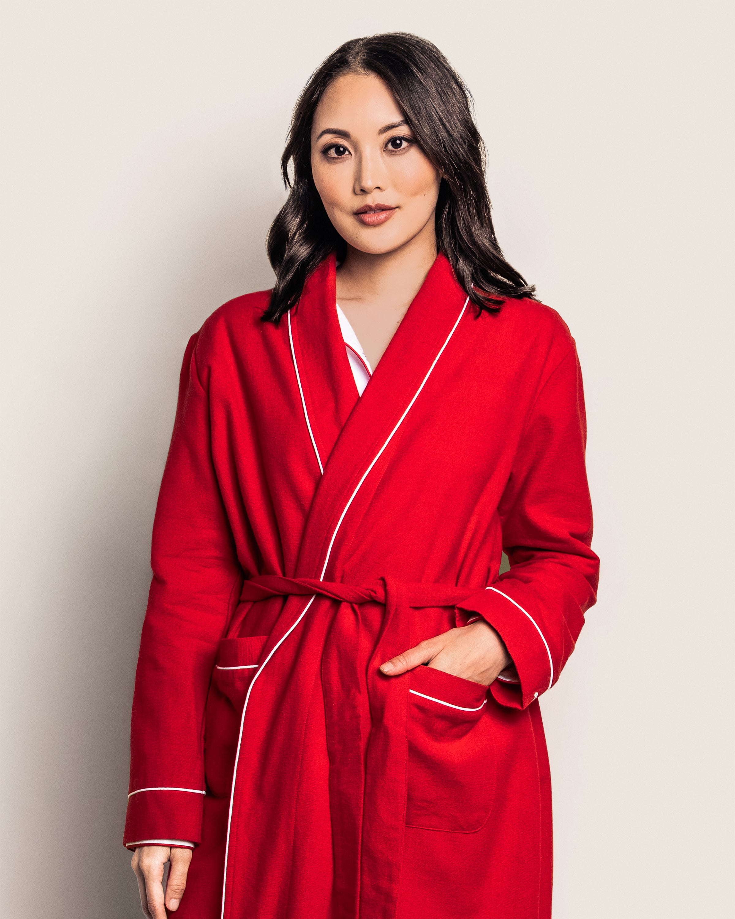 A woman in a Petite Plume Womens Flannel Robe in Red with White Piping, featuring subtle monogramming, stands against a light background. Her shoulder-length dark hair and relaxed expression enhance the cozy vibe, with one hand casually tucked into the robes pocket.