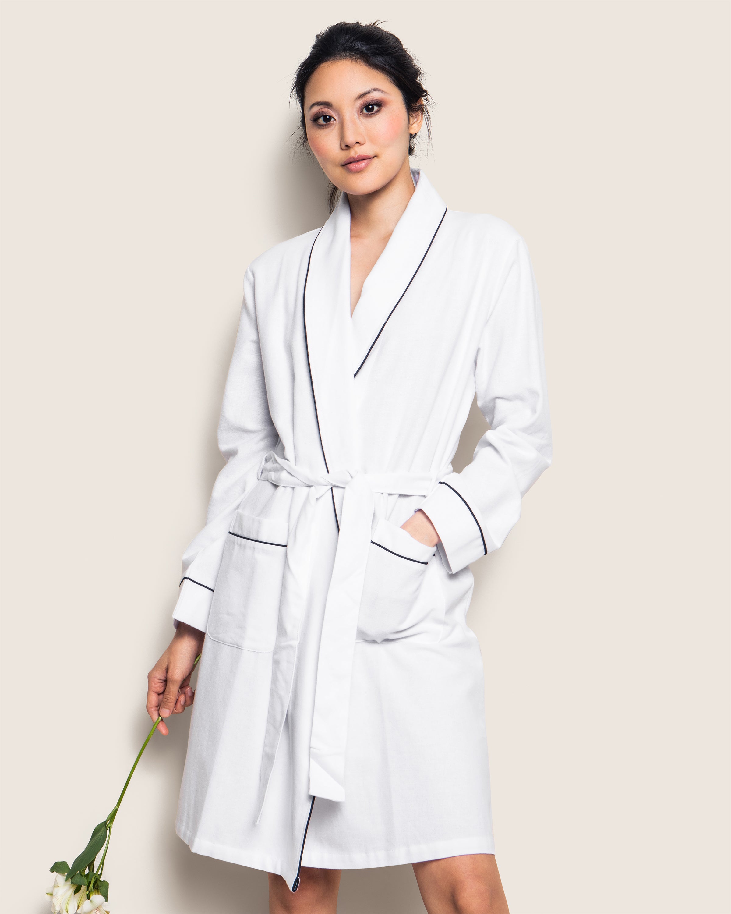 Against a neutral backdrop, a person in Petite Plumes Womens Flannel Robe in White with Navy Piping gracefully holds a white flower. Made from premium cotton, the robe is belted at the waist with hands resting gently in its pockets.