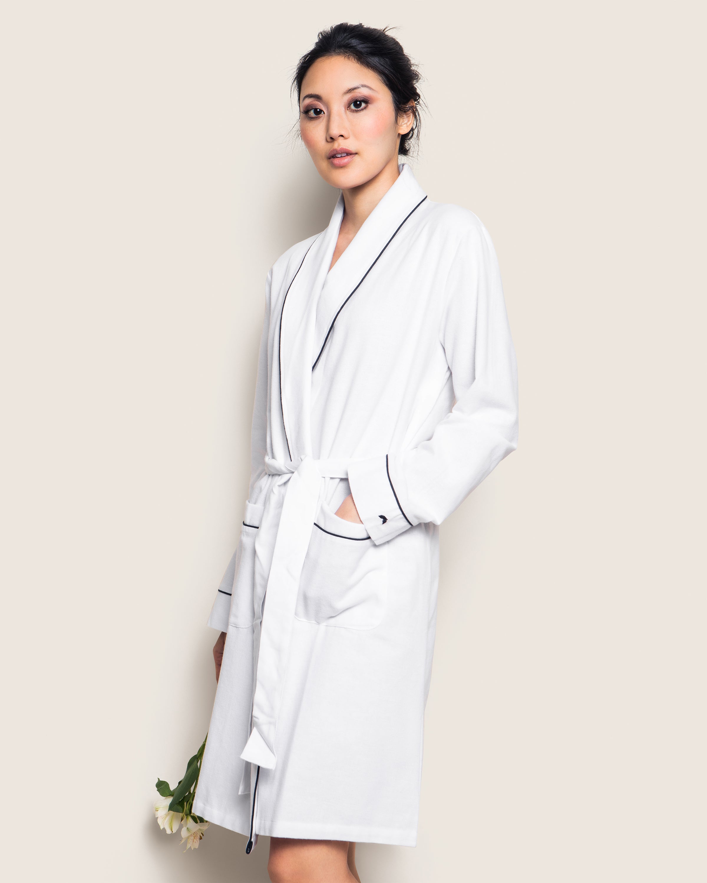Standing against a neutral background, a person wears the Petite Plume Womens Flannel Robe in White with Navy Piping. Made from premium cotton, it features two front pockets and is tied at the waist. They hold a flower, looking calm and relaxed.