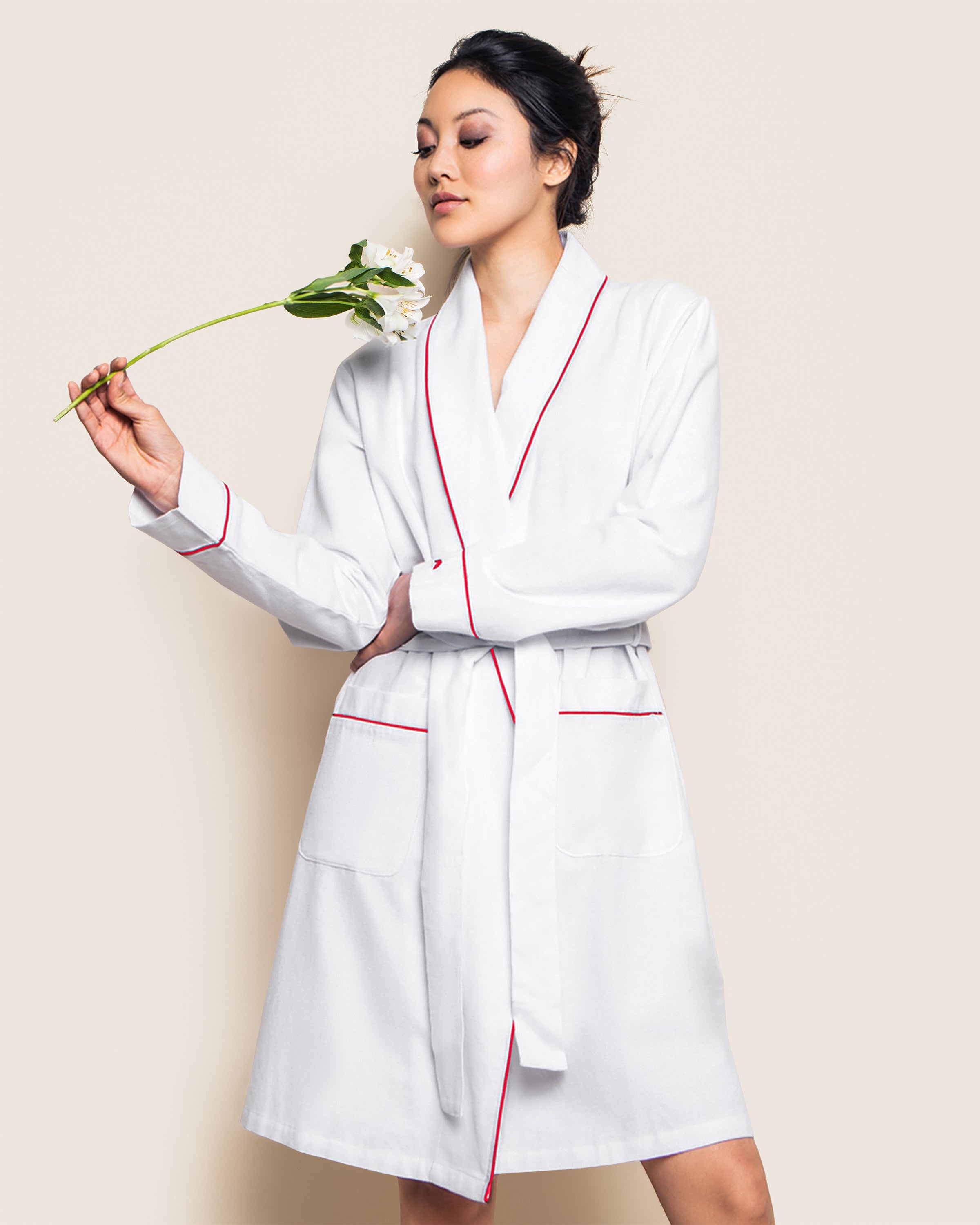 A person is wearing a Petite Plume Womens Flannel Robe in White with Red Piping, holding a white flower with closed eyes against a plain, light background.