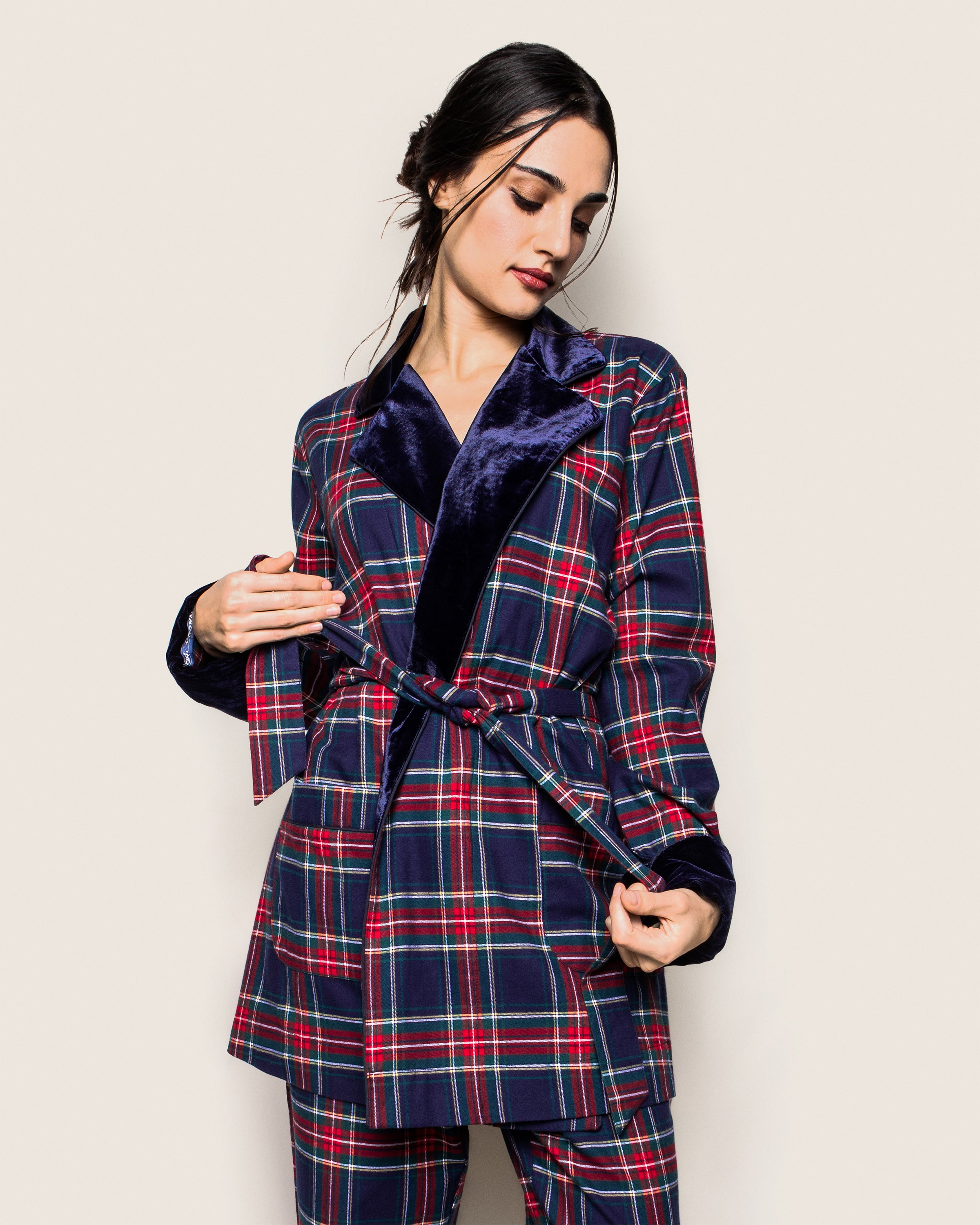 A woman with dark hair pulled back is against a light neutral background, adjusting the belt of her Petite Plume Womens Car Coat with Velvet Trim in Windsor Tartan. The plaid design has red, blue, and white patterns on lightweight flannel weave for elegance as she looks down to her right.