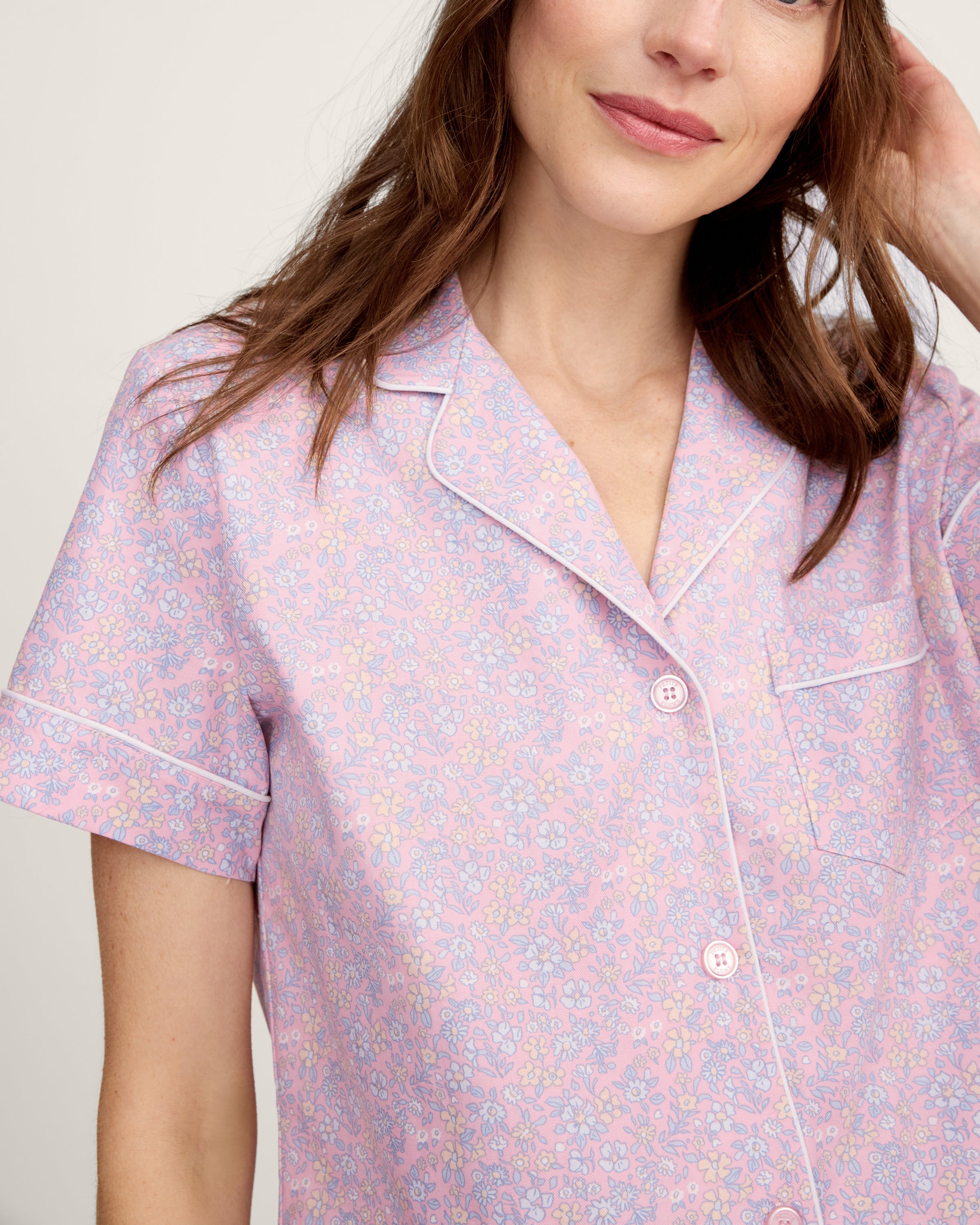 A woman with long brown hair is wearing the Women's Twill Short Sleeve Short Set in Fleur Blush and smiling softly, standing in front of a plain light background.