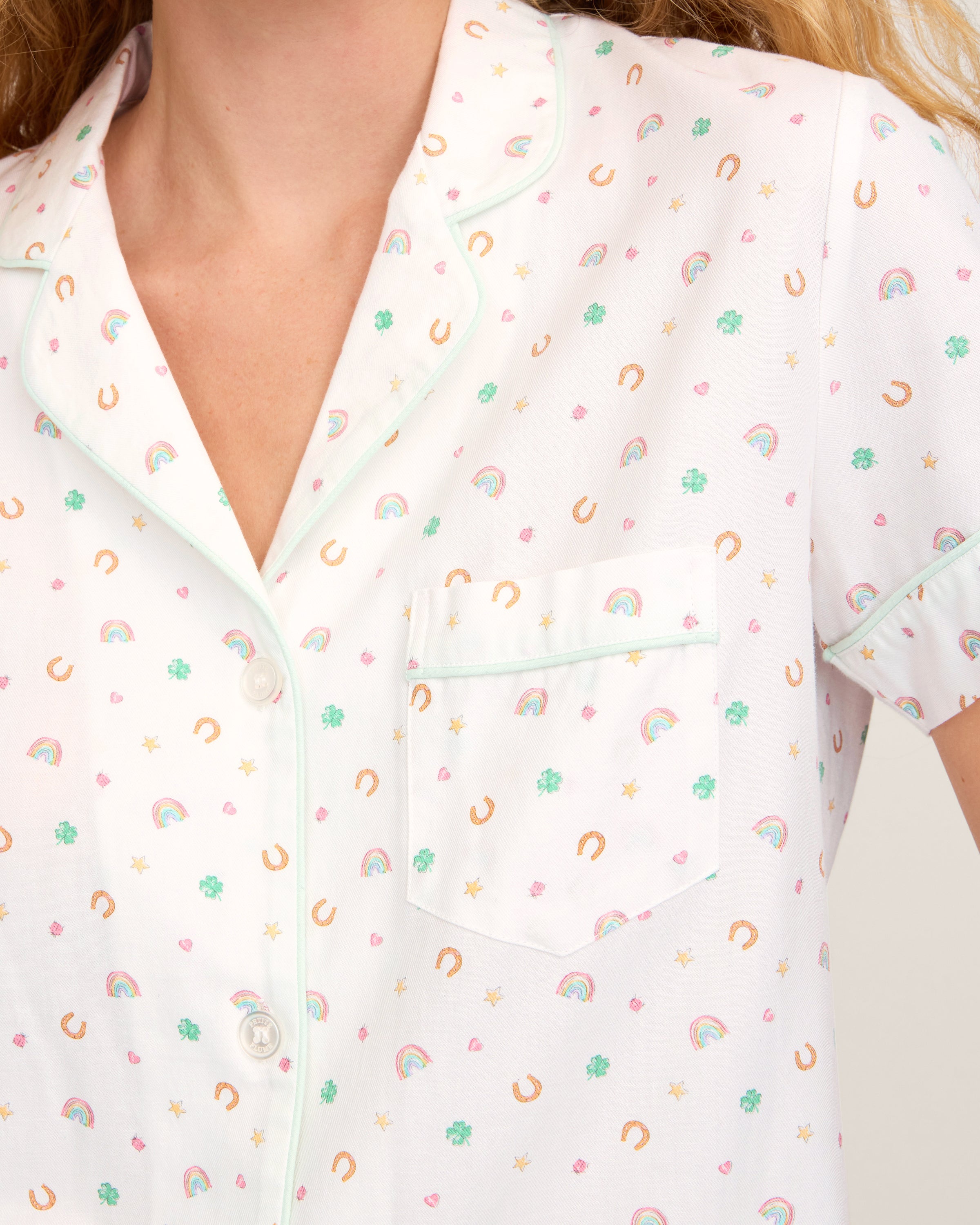 A woman with long, wavy blonde hair wears the Women's Twill Short Sleeve Short Set in Good Luck Charms, featuring a multicolored pattern, standing against a plain light background.