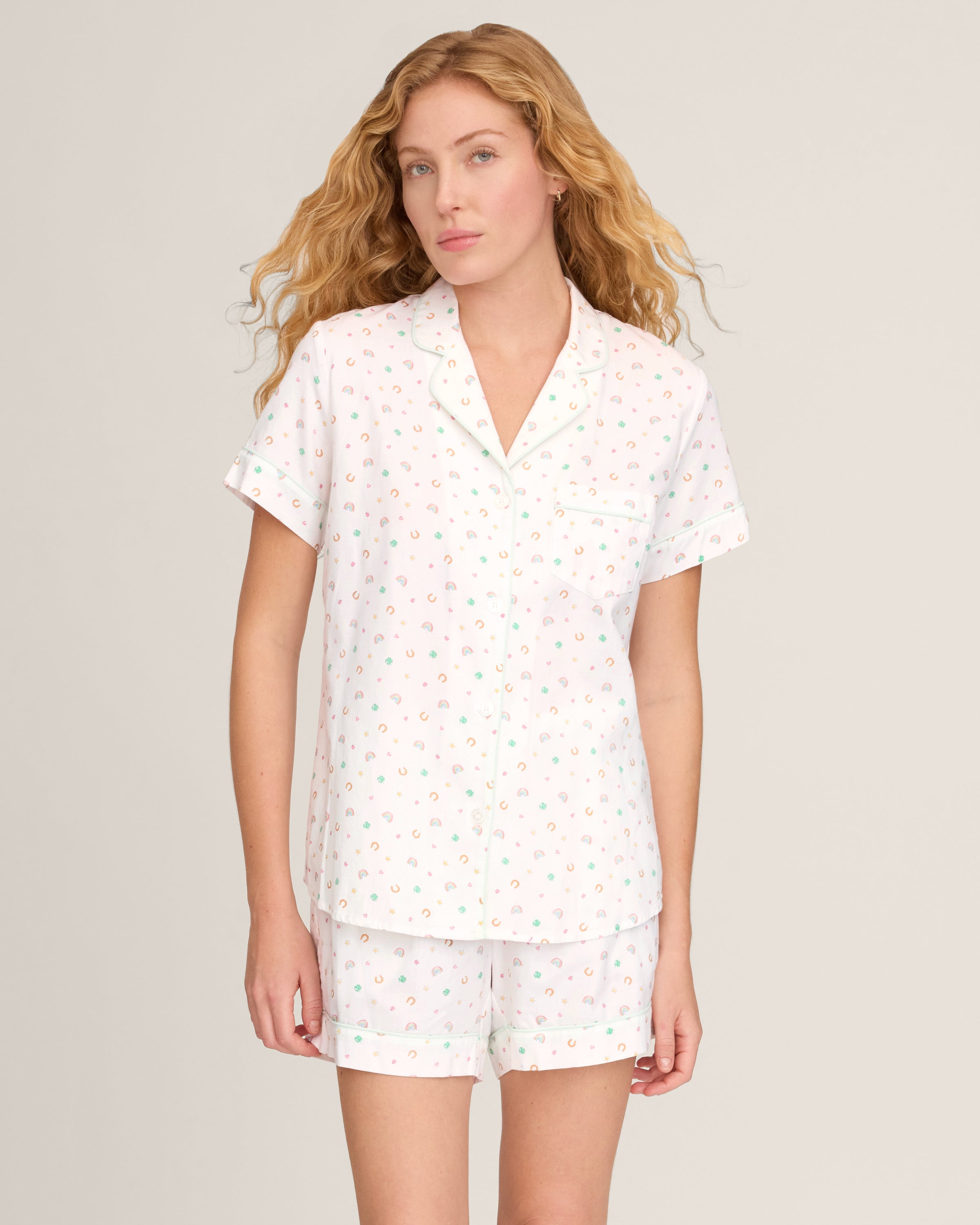 A woman with long, wavy blonde hair wears the Women's Twill Short Sleeve Short Set in Good Luck Charms, featuring a multicolored pattern, standing against a plain light background.