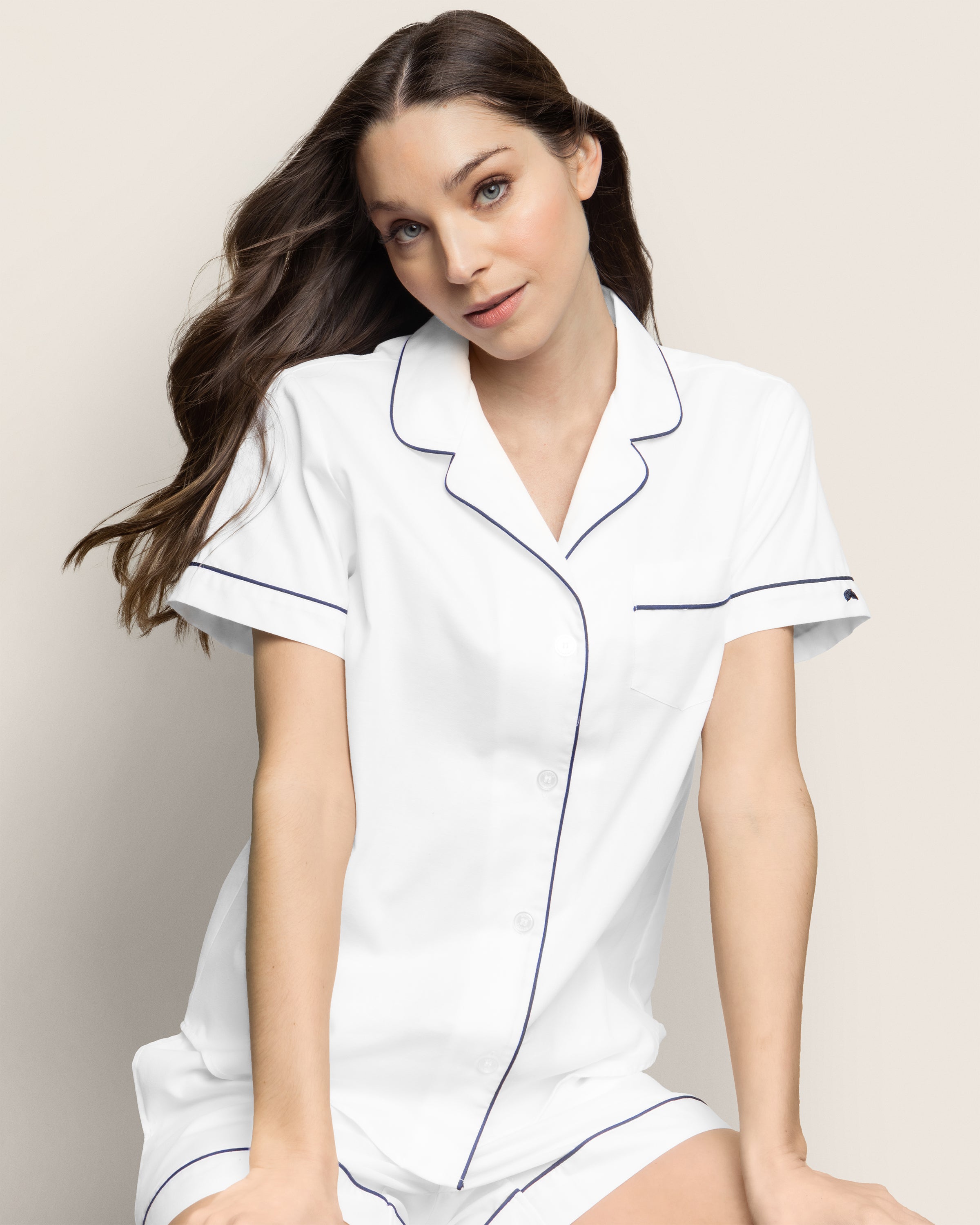 A woman with long brown hair sits gracefully wearing the Womens Twill Pajama Short Set in White with Navy Piping by Petite Plume. Her white button-up pajama set exudes timeless elegance against the soft, neutral backdrop, epitomizing luxurious sleepwear.