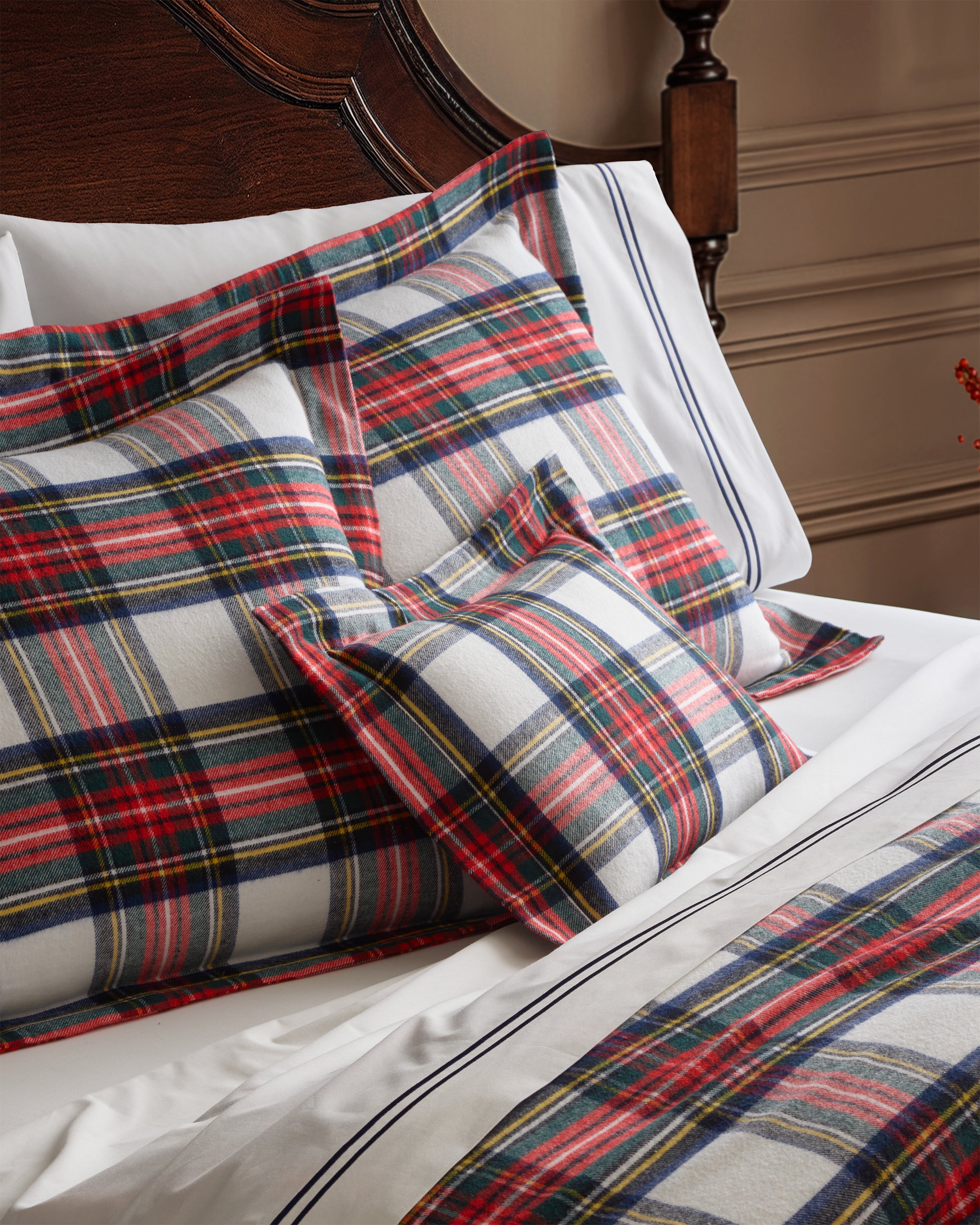 The bed is adorned with Petite Plumes Balmoral Tartan pillows in red, blue, and white plaid. These flannel decorative accents add a charming holiday flair against the dark wooden headboard and beige walls.