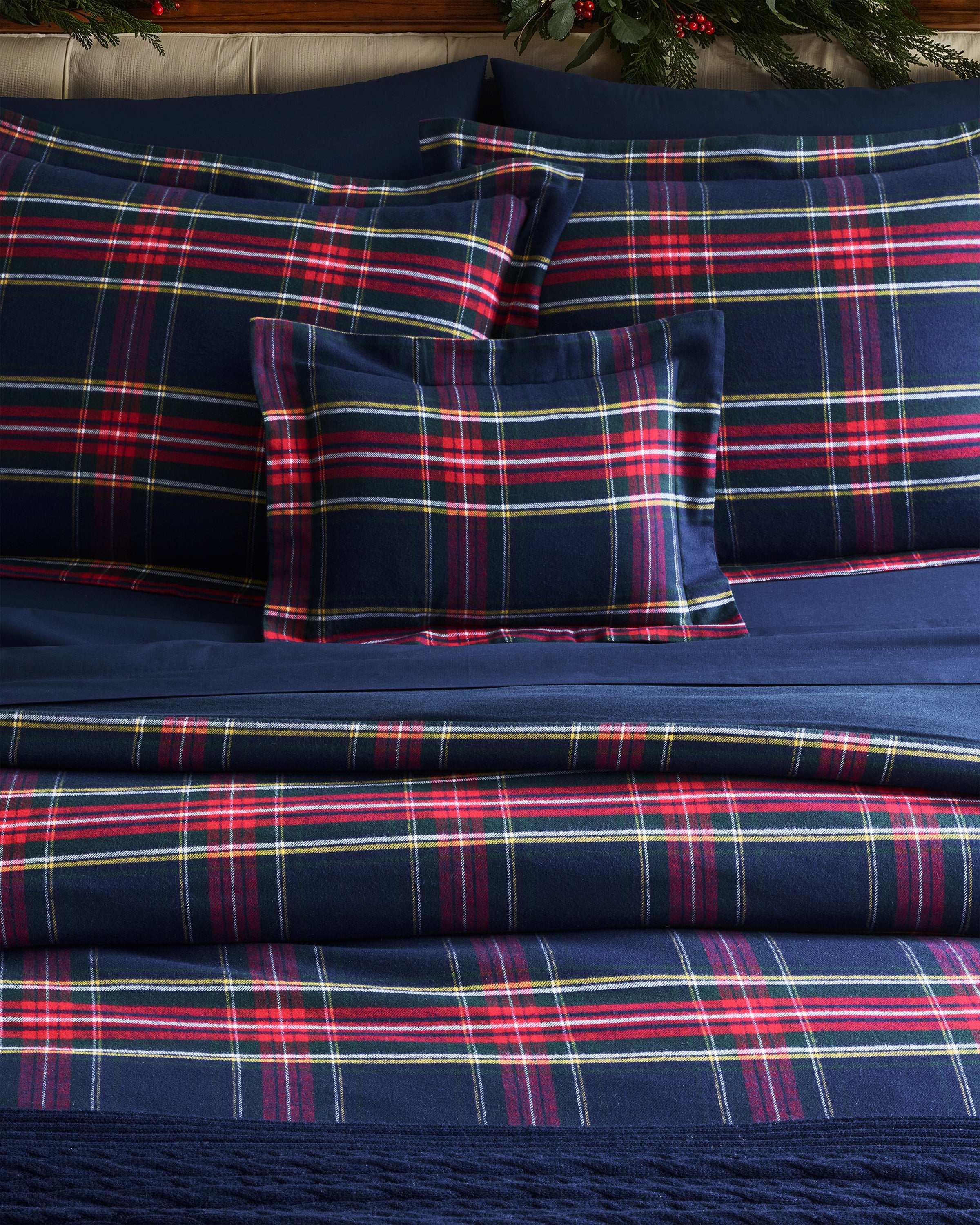 A bed featuring Petite Plumes Windsor Tartan bedding in brushed cotton flannel displays classic blue, red, and white plaid prints. Neatly arranged pillows and comforter enhance intersecting stripes, while greenery with red berries adds charm to the headboard area.