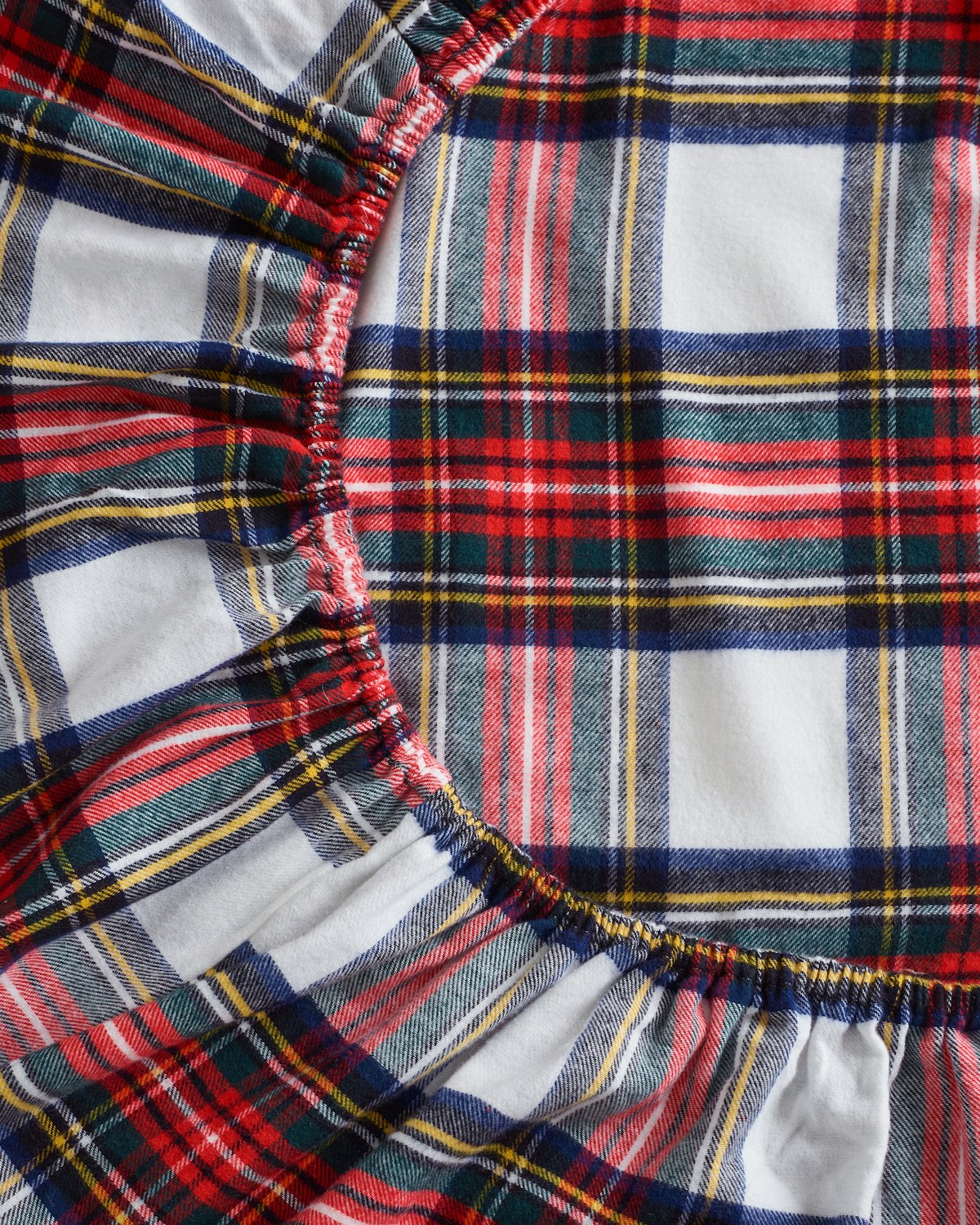 Close-up of Petite Plumes Flannel Crib Sheet in Balmoral Tartan showcasing a vibrant plaid design with red, white, blue, green, and yellow stripes. The circular gathered pattern creates a ruffled texture similar to a cozy cotton flannel sheet set.