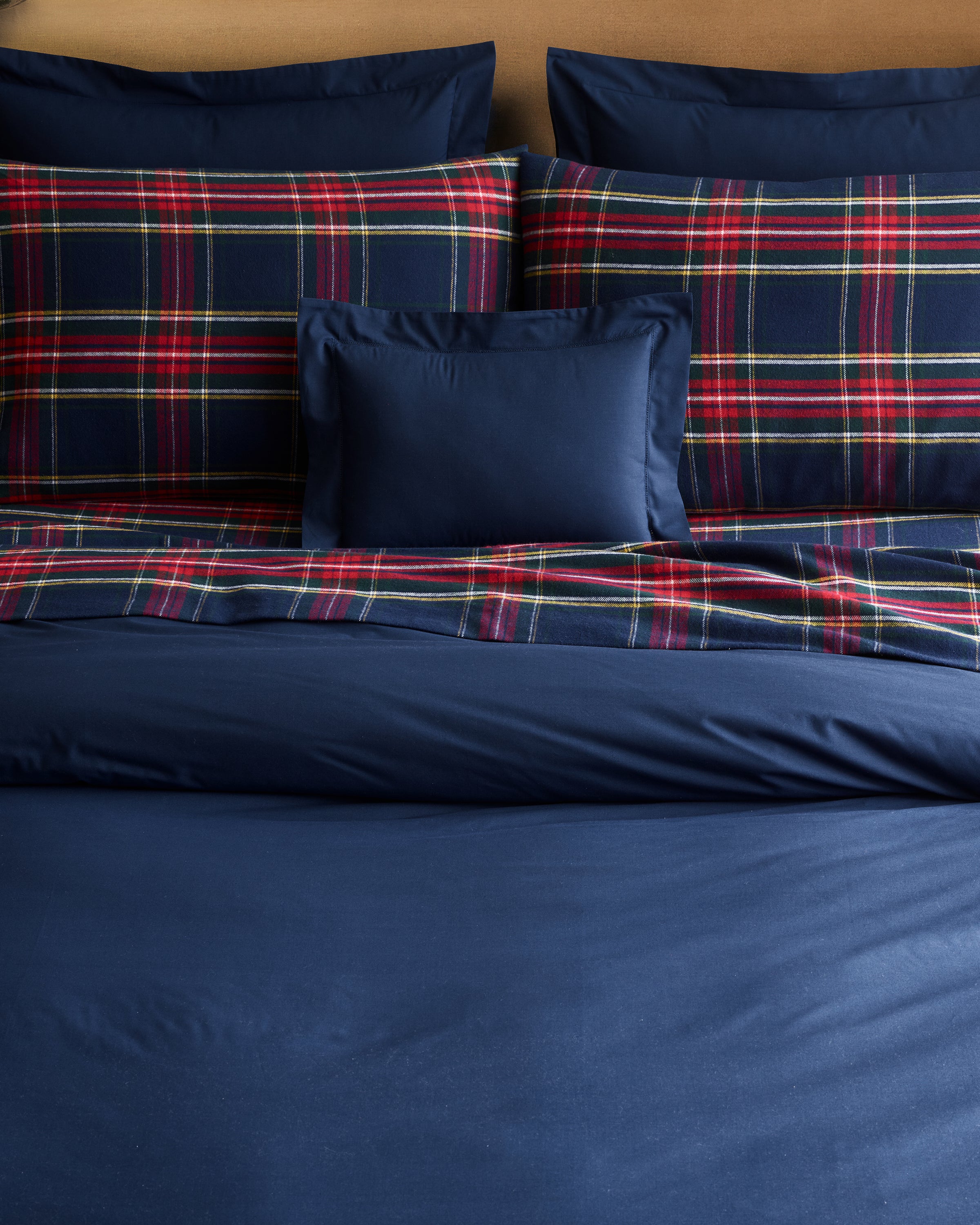 A neatly made bed showcases Petite Plumes Flannel Sheets in Windsor Tartan, featuring navy sheets and pillowcases. Two large pillows and a matching red and green plaid blanket ensure cozy warmth, while a smaller navy rectangular pillow centers the look.