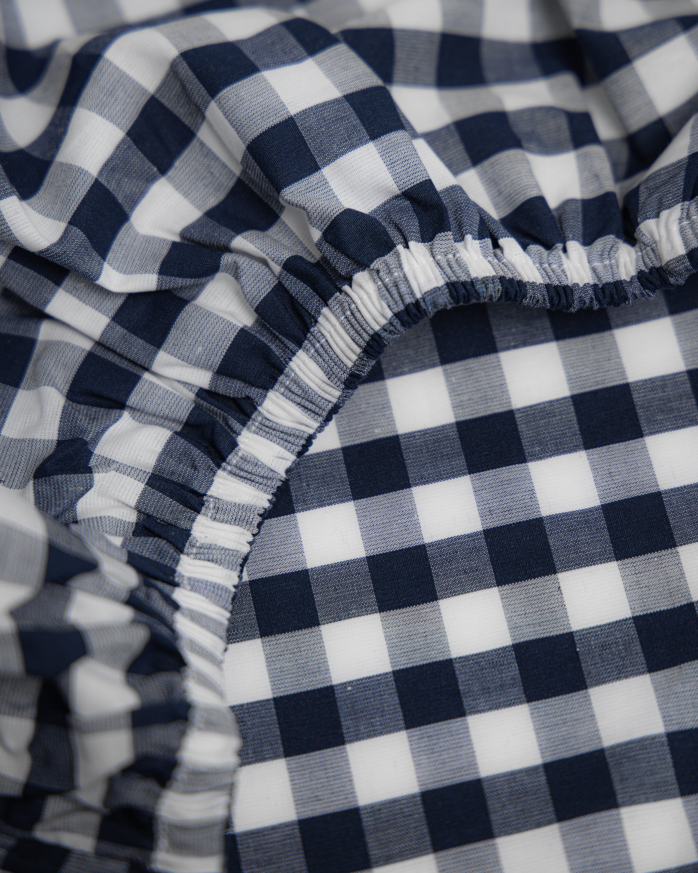 Featuring a close-up of the ultra-soft Luxe Premium Cotton Navy Gingham Crib Sheet by Petite Plume, this elastic-gathered sheet shows creases and ruffles. Its prominent texture highlights the classic navy gingham check pattern.
