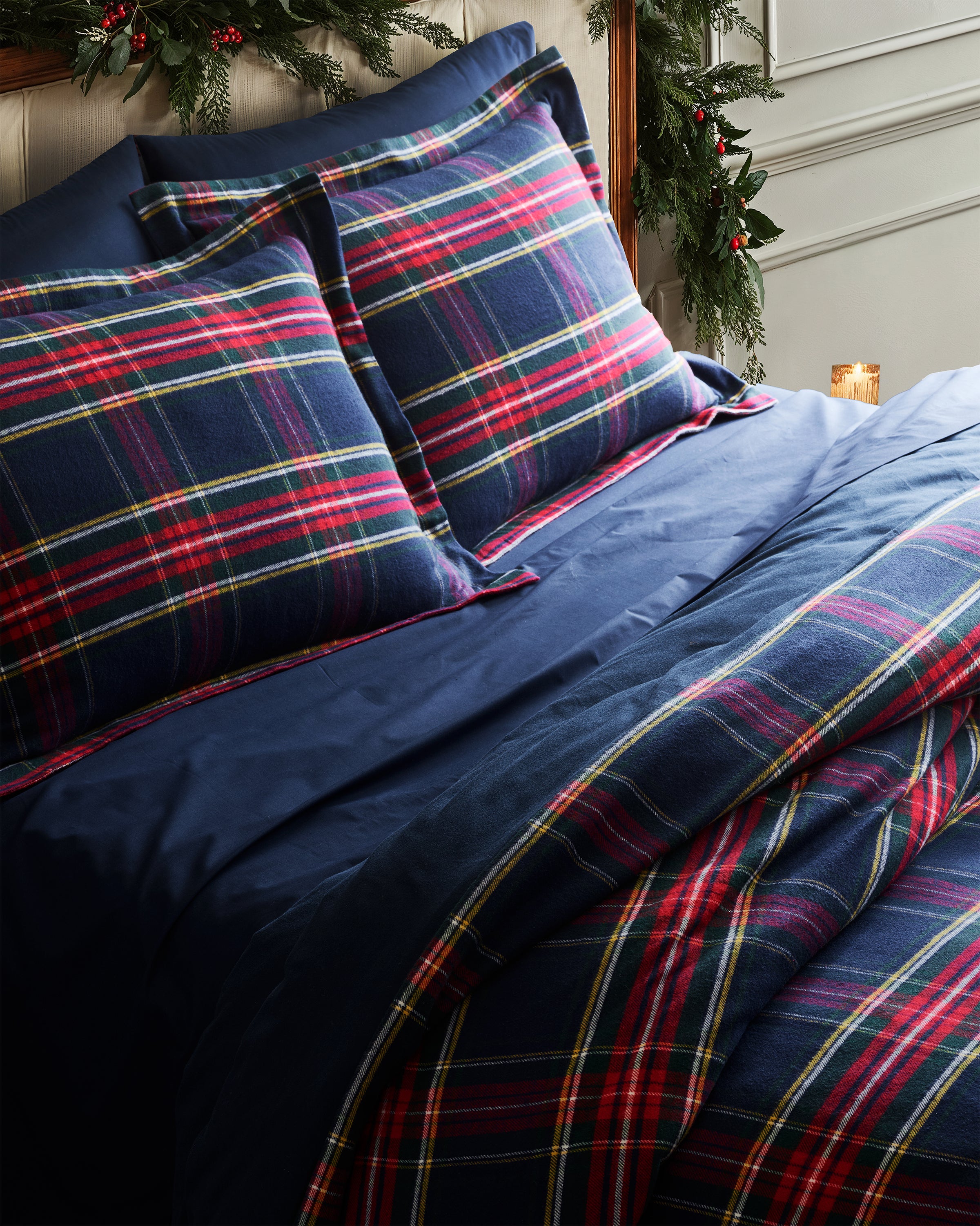 A cozy bed with Luxe Premium Cotton Bed Sheets in Navy by Petite Plume, accented with green garland and a lit candle on the bedside table, creates a festive atmosphere.