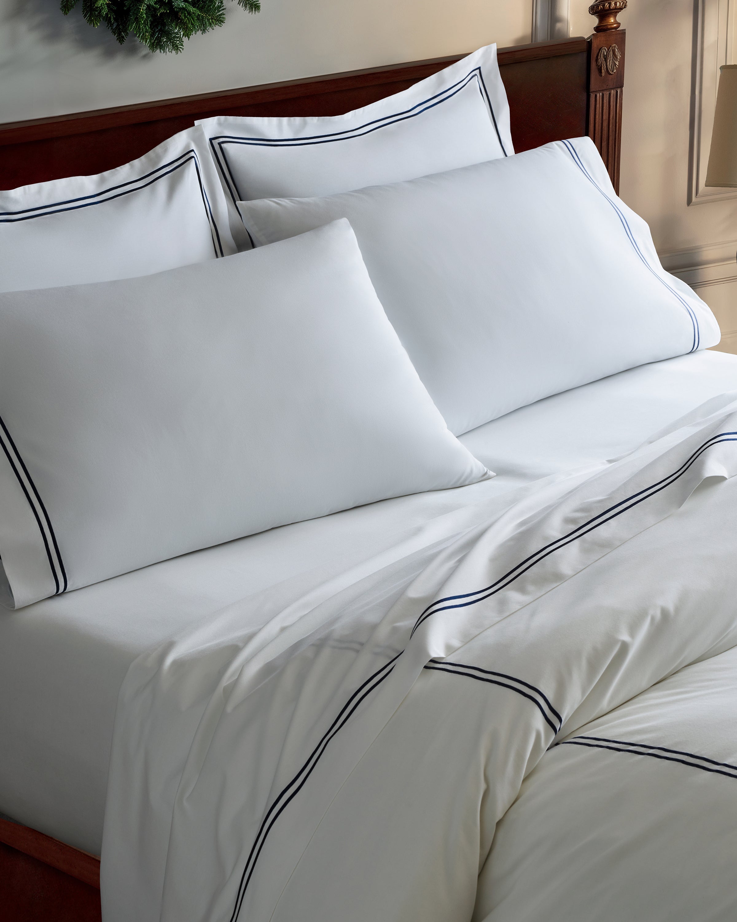 The bed showcases Petite Plumes Luxe Premium Cotton Sheets in White with Navy trim. The long-staple cotton sheets and pillowcases are neatly arranged, featuring two large pillows against the headboard and a comforter folded at the foot of the bed for a cozy look.