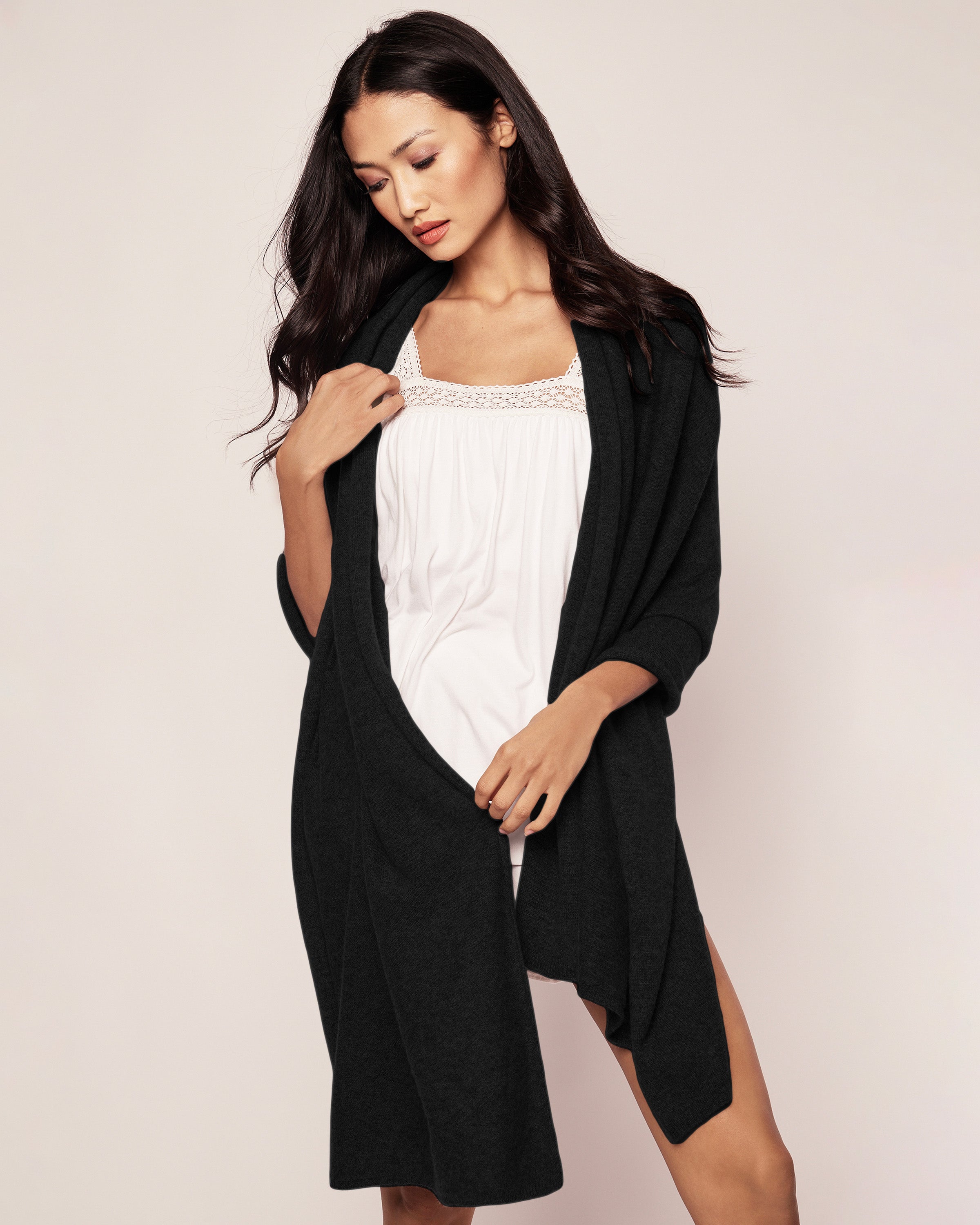A woman wearing Petite Plumes Womens Cashmere Wrap in Black over a white dress stands against a plain background. She looks down slightly, adjusting the wrap, which elegantly drapes around her shoulders.