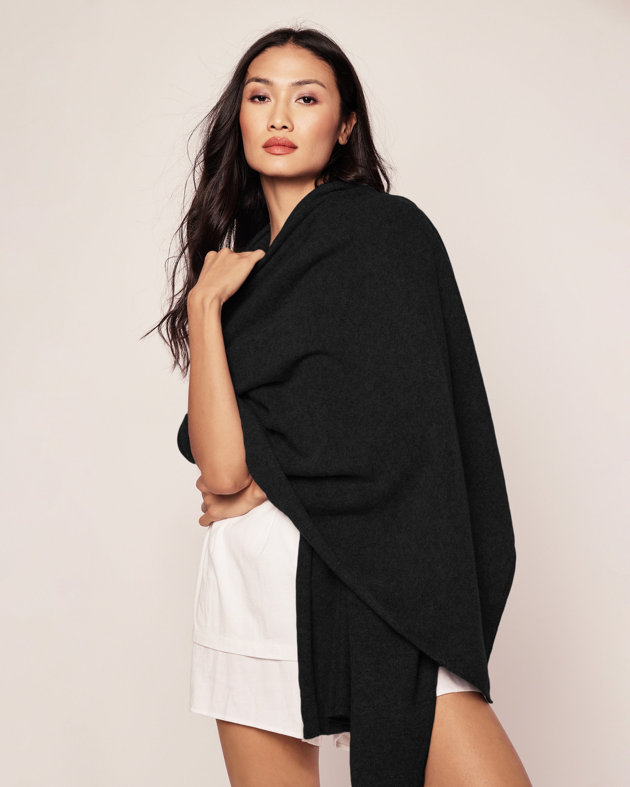 A woman with long dark hair, wearing a white outfit and a large Petite Plume Womens Cashmere Wrap in Black draped elegantly around her shoulders, stands confidently against a plain background, looking toward the camera.
