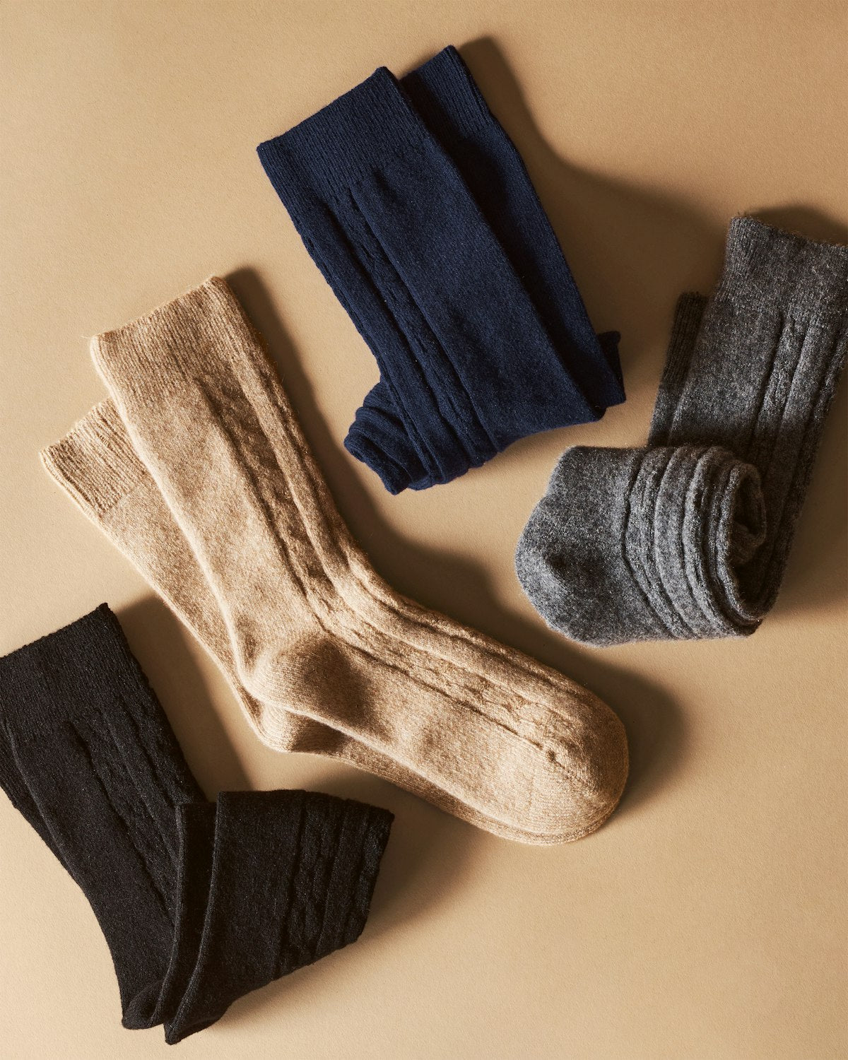 Four pairs of luxurious Womens Cashmere Socks by Petite Plume, crafted from Grade A cashmere in black, beige, navy, and gray, are displayed on a beige background. The socks are either neatly folded or laid flat.