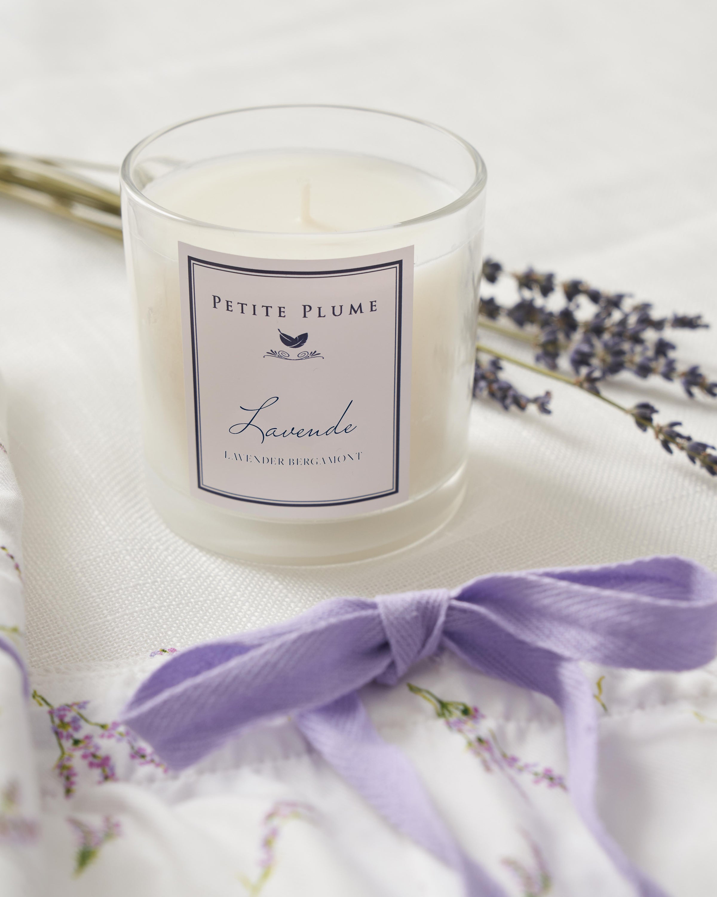 A Petite Plume Lavande Candle from the Petite Plume collection elegantly sits in a glass jar on a white surface, surrounded by sprigs of lavender and a matching ribbon to complete the tranquil aesthetic.