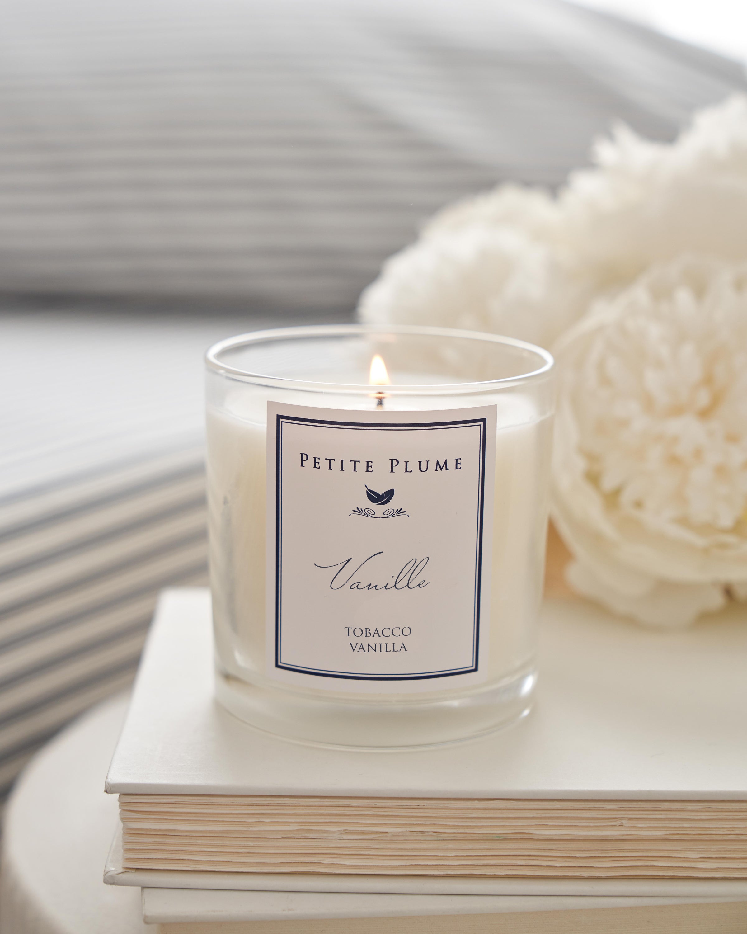 A lit Petite Plume Luxe Vanille Candle from the Candle Collection rests on a stack of white books, with softly blurred white flowers and a bed featuring striped bedding in the background, enhancing the serene ambiance.