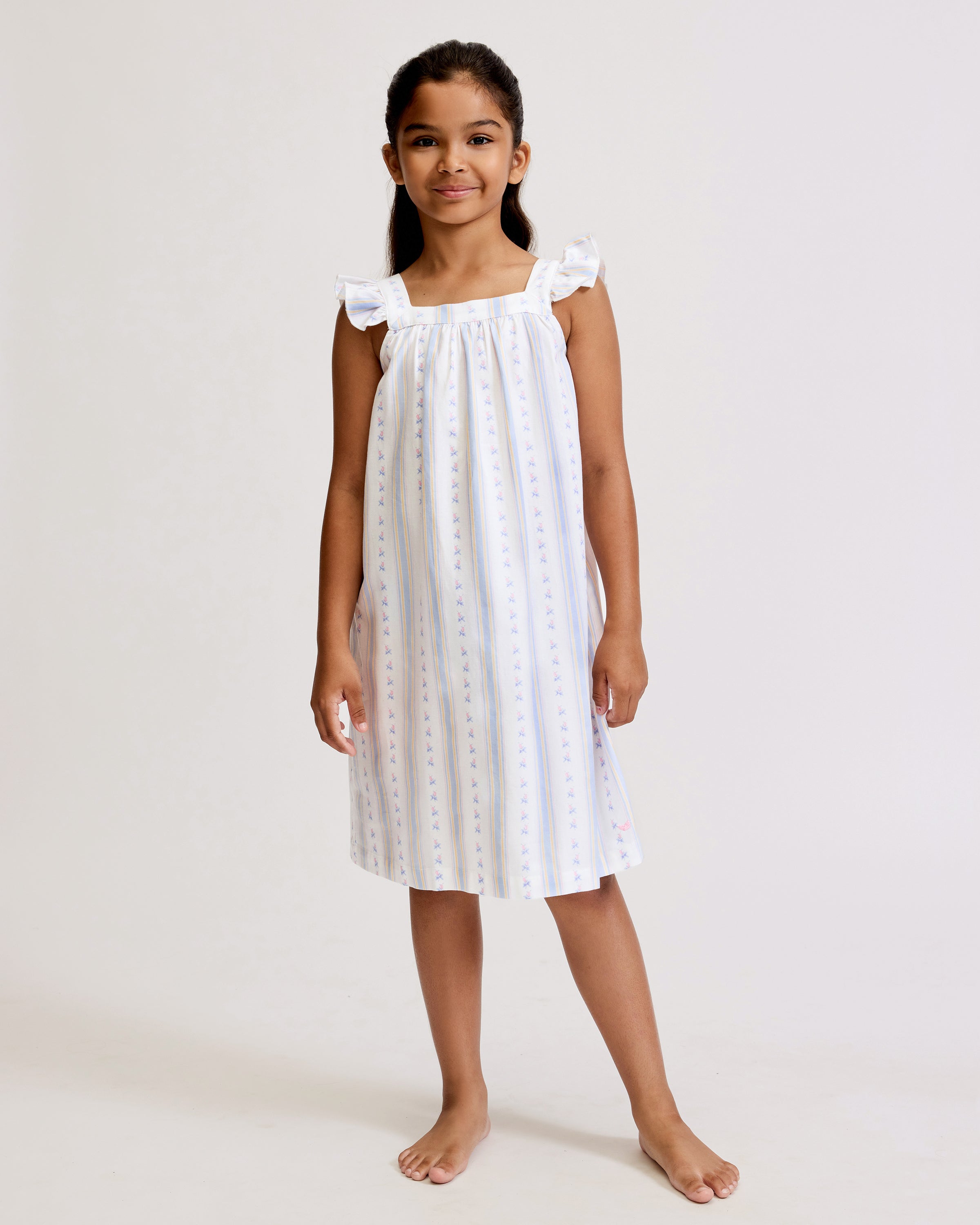 A young girl with long dark hair stands barefoot, smiling, wearing the Girl's Twill Clara Nightgown in Cottage Stripe—white with subtle pastel stripes and ruffled shoulders—against a plain light background.