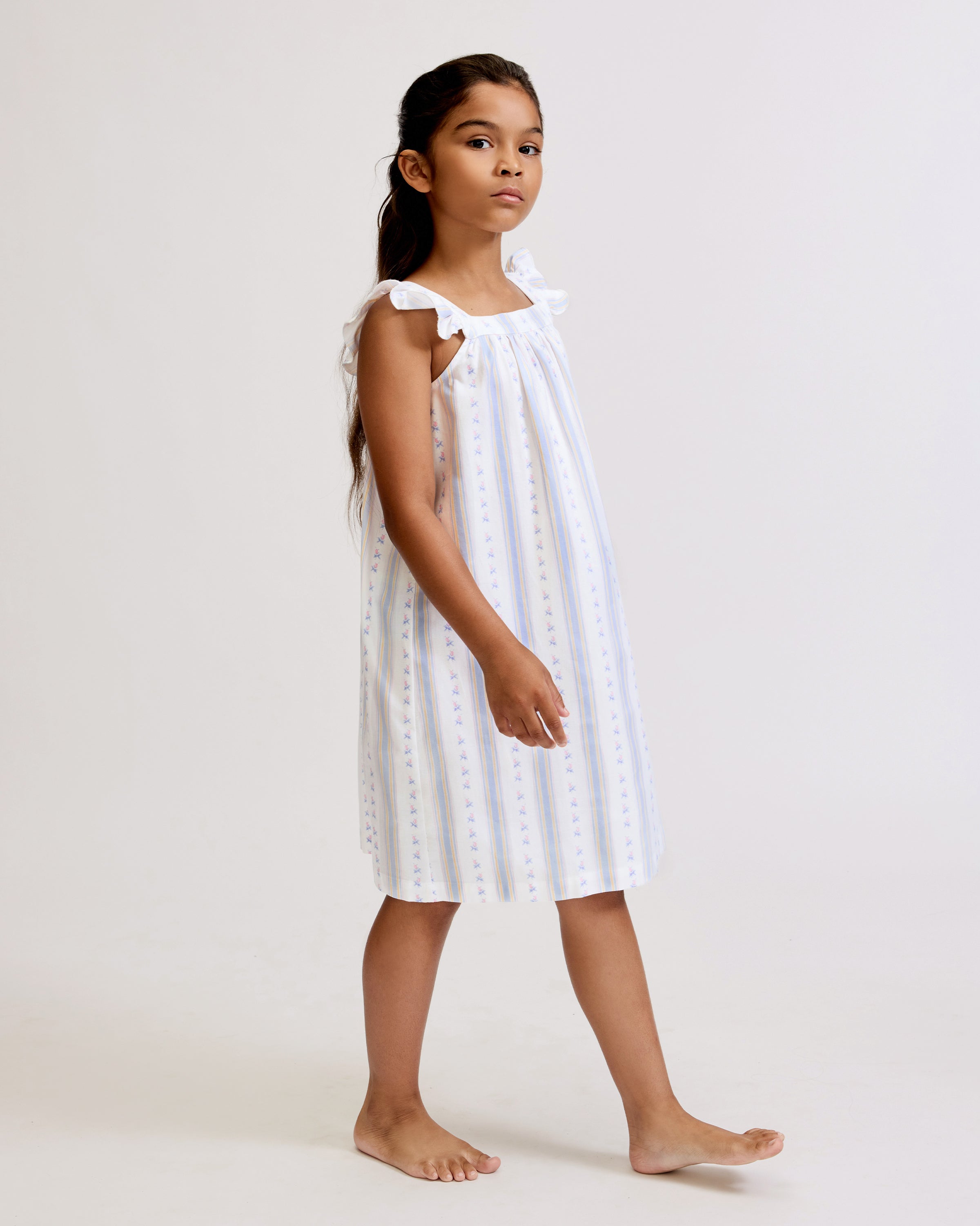 A young girl with long dark hair stands barefoot, smiling, wearing the Girl's Twill Clara Nightgown in Cottage Stripe—white with subtle pastel stripes and ruffled shoulders—against a plain light background.