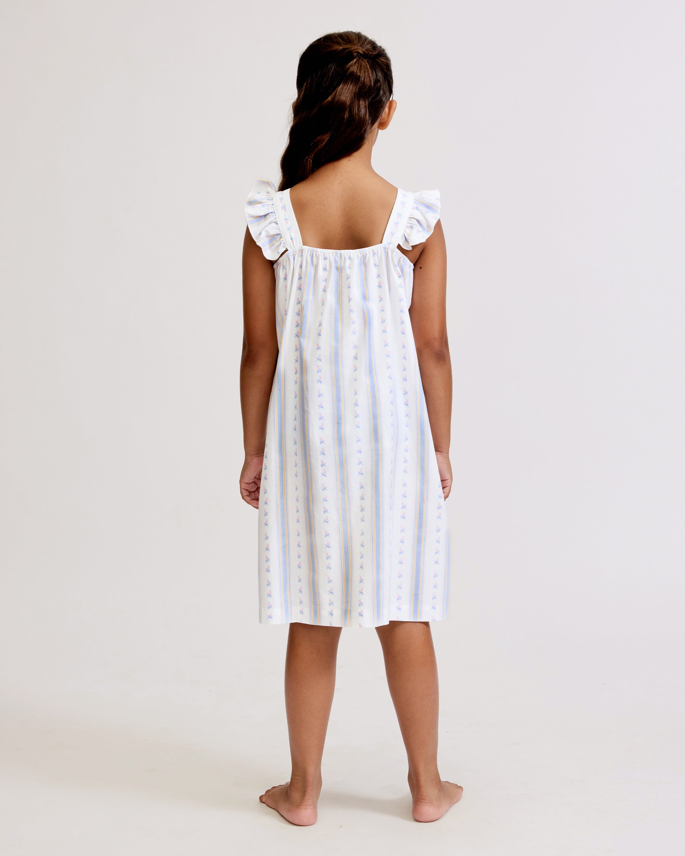 A young girl with long dark hair stands barefoot, smiling, wearing the Girl's Twill Clara Nightgown in Cottage Stripe—white with subtle pastel stripes and ruffled shoulders—against a plain light background.