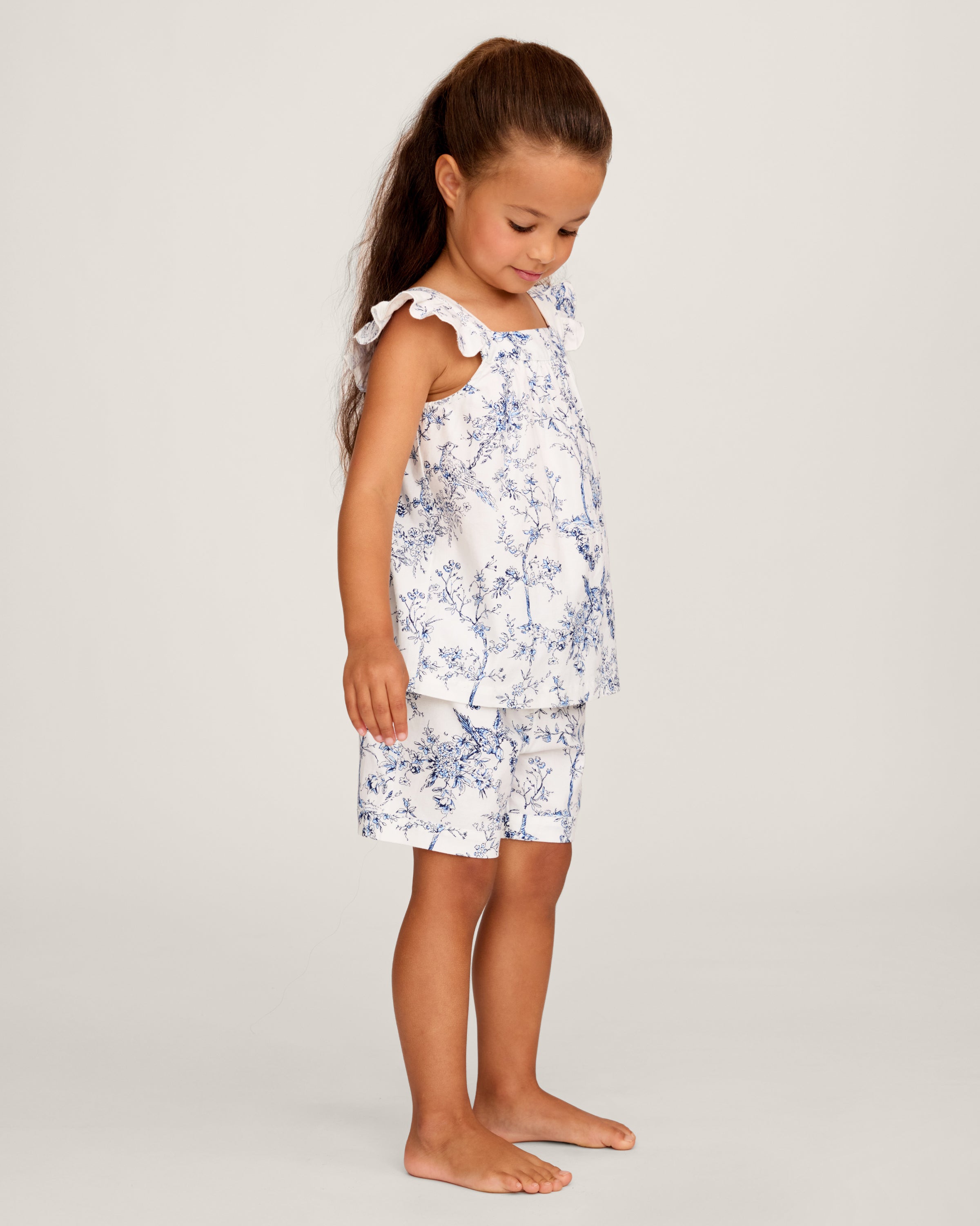 A young girl with long brown hair stands barefoot, smiling in the Girl's Twill Clara Short Set in Timeless Toile—a white top and matching blue floral shorts—posing against a plain light background.
