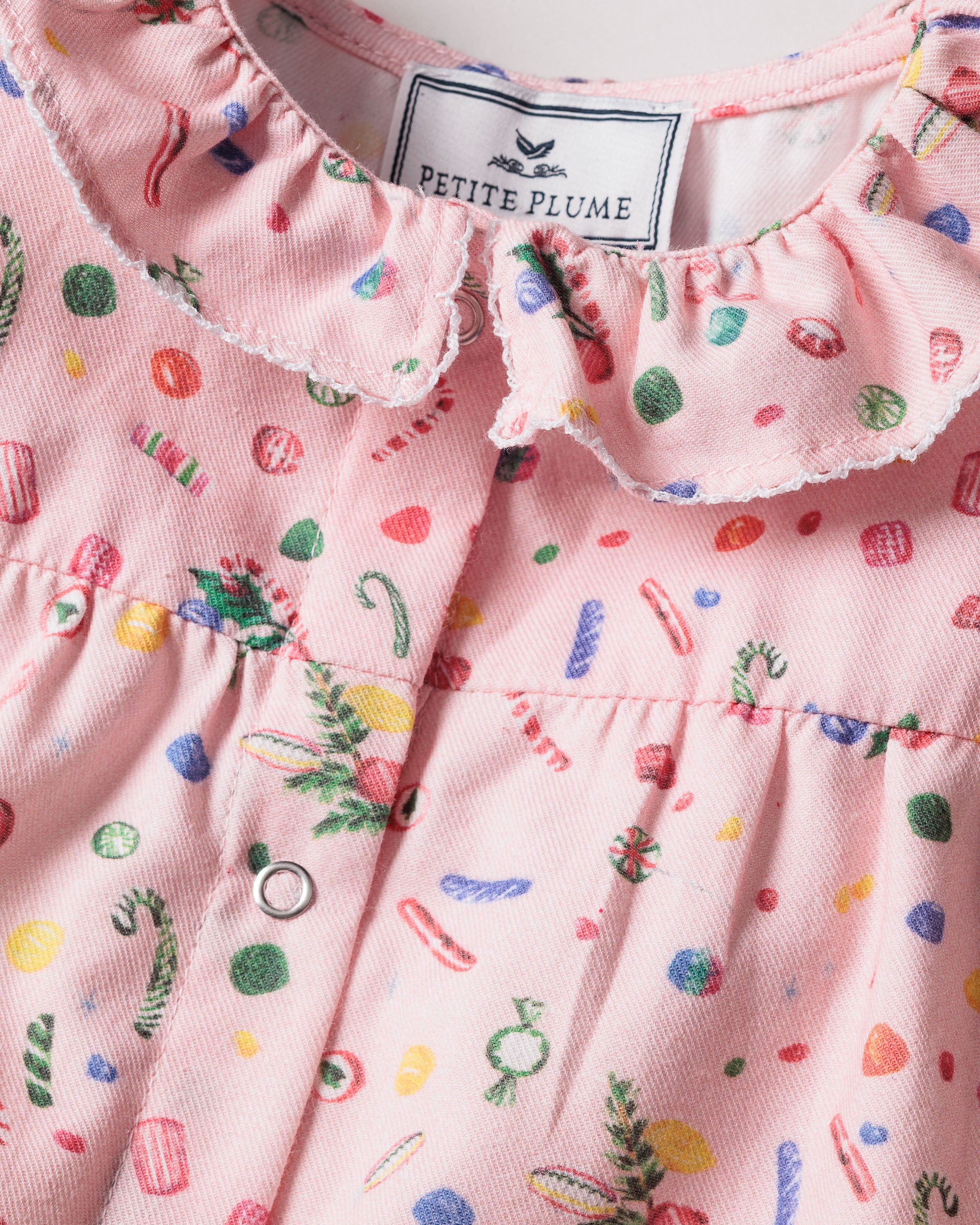 The Babys Twill Madeline Romper in Vintage Sweets by Petite Plume features a pink fabric with a colorful candy print, ideal for holiday dreams. It has a ruffled collar and snap buttons down the front.