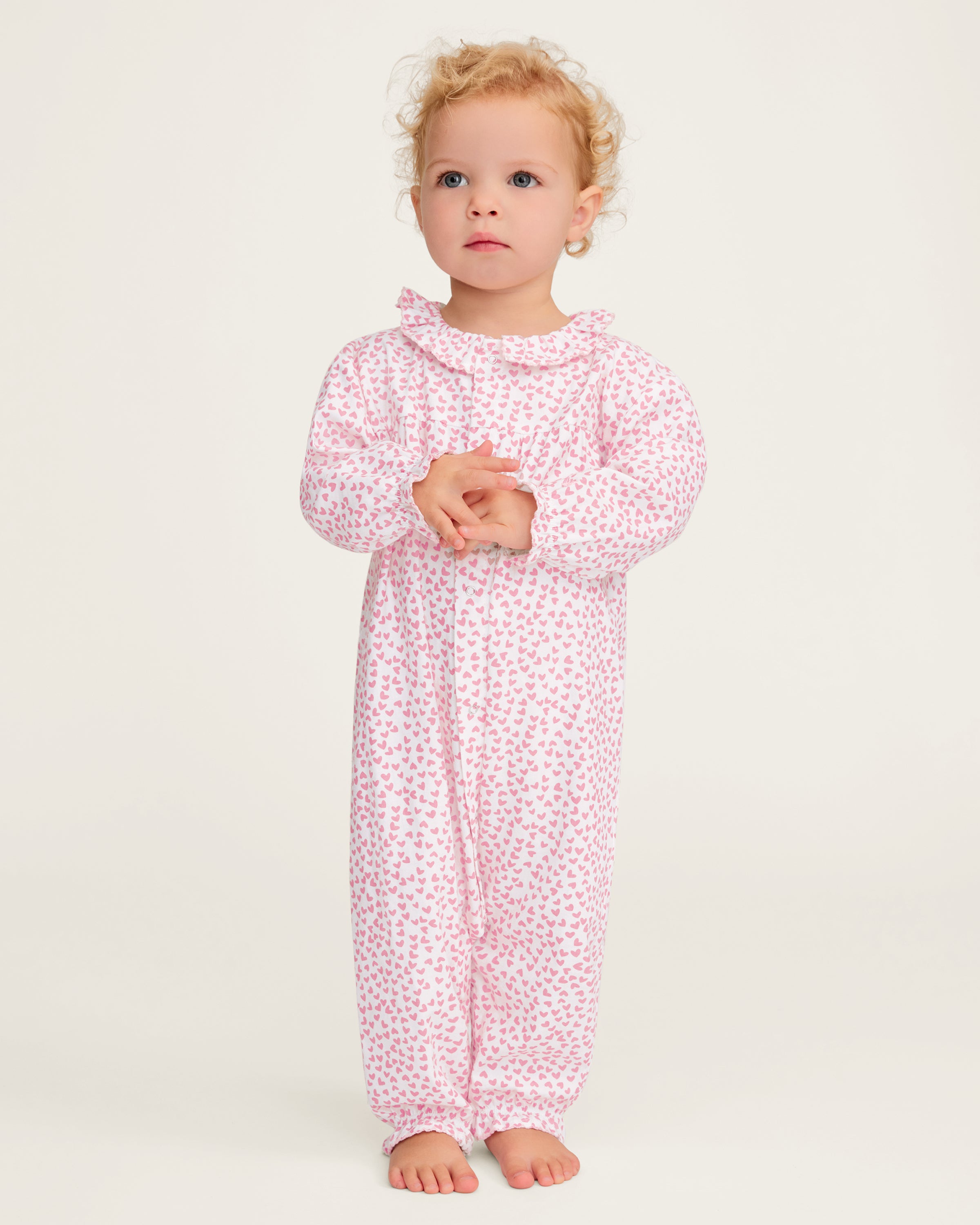 The Baby's Twill Madeline Romper in Sweethearts is a pink and white long-sleeve romper featuring a ruffled collar and cuffs, adorned with a small pink heart pattern.