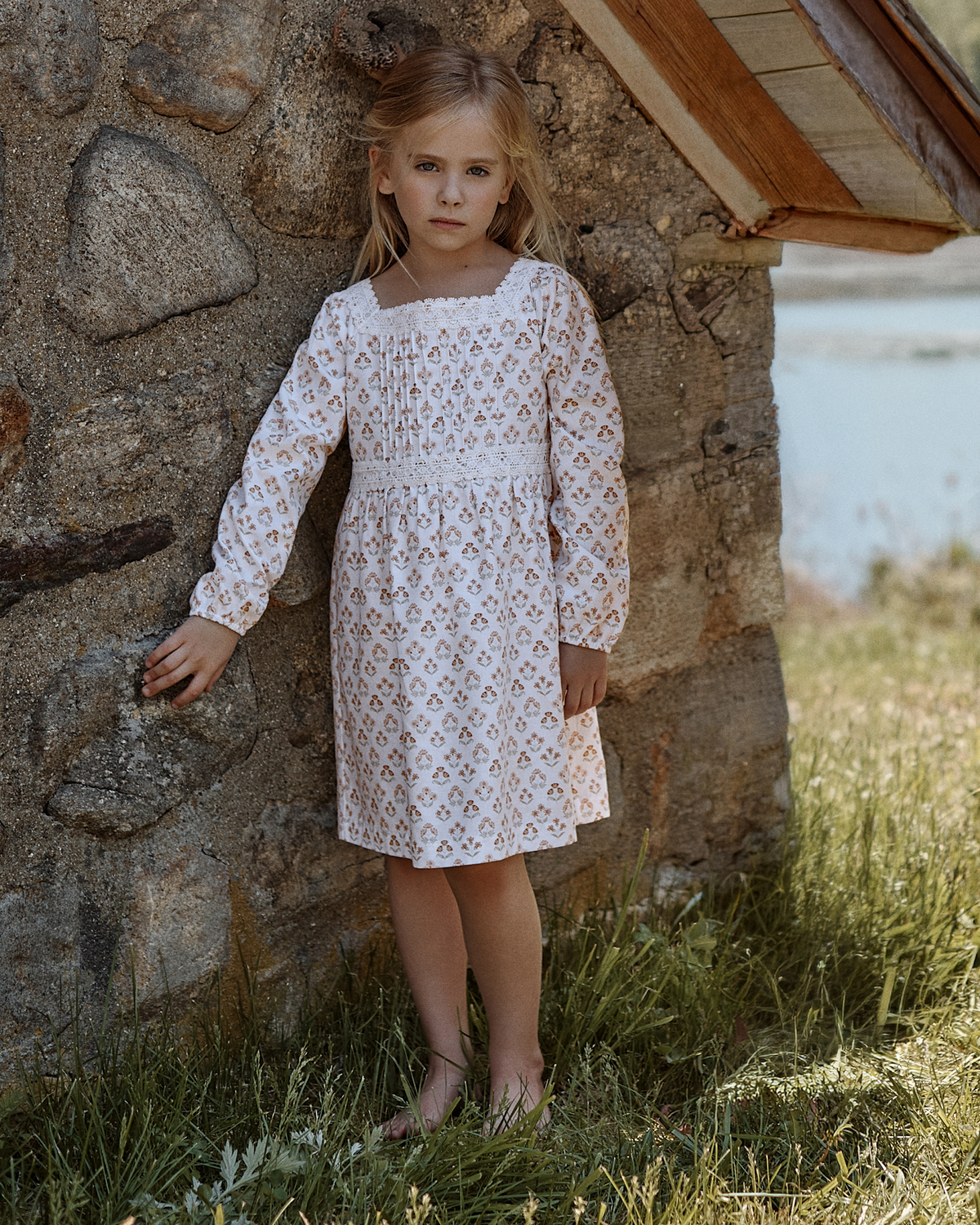A young girl stands barefoot by a stone wall outside, near grass and water, wearing the Girl’s Twill Long Sleeve Juliette Nightgown in Estate Garden and looking seriously at the camera.