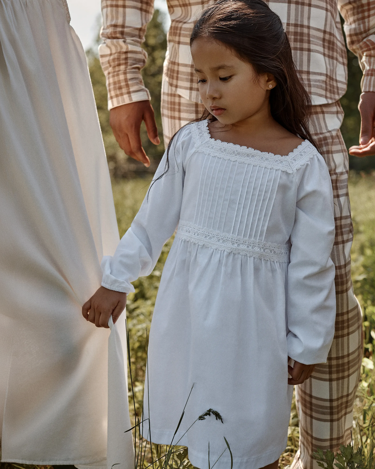 A young girl wearing the Girl’s Twill Long Sleeve Juliette Nightgown in White stands outdoors, holding the hand of an adult beside her. Two adults dressed in white and plaid are partially visible in the background, surrounded by greenery.