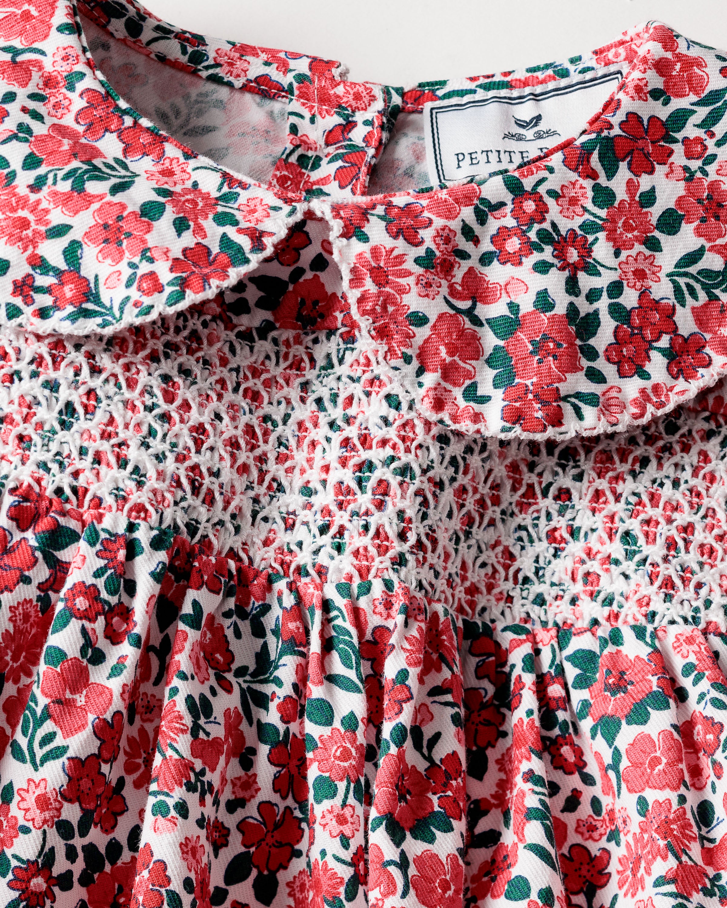 Close-up of Petite Plumes Girls Eloise Nightgown in Fleurs des Bois, featuring red flowers and green leaves on white. It has a round collar and smocked chest details, with a visible label for comfort in this moisture-wicking twill weave garment.